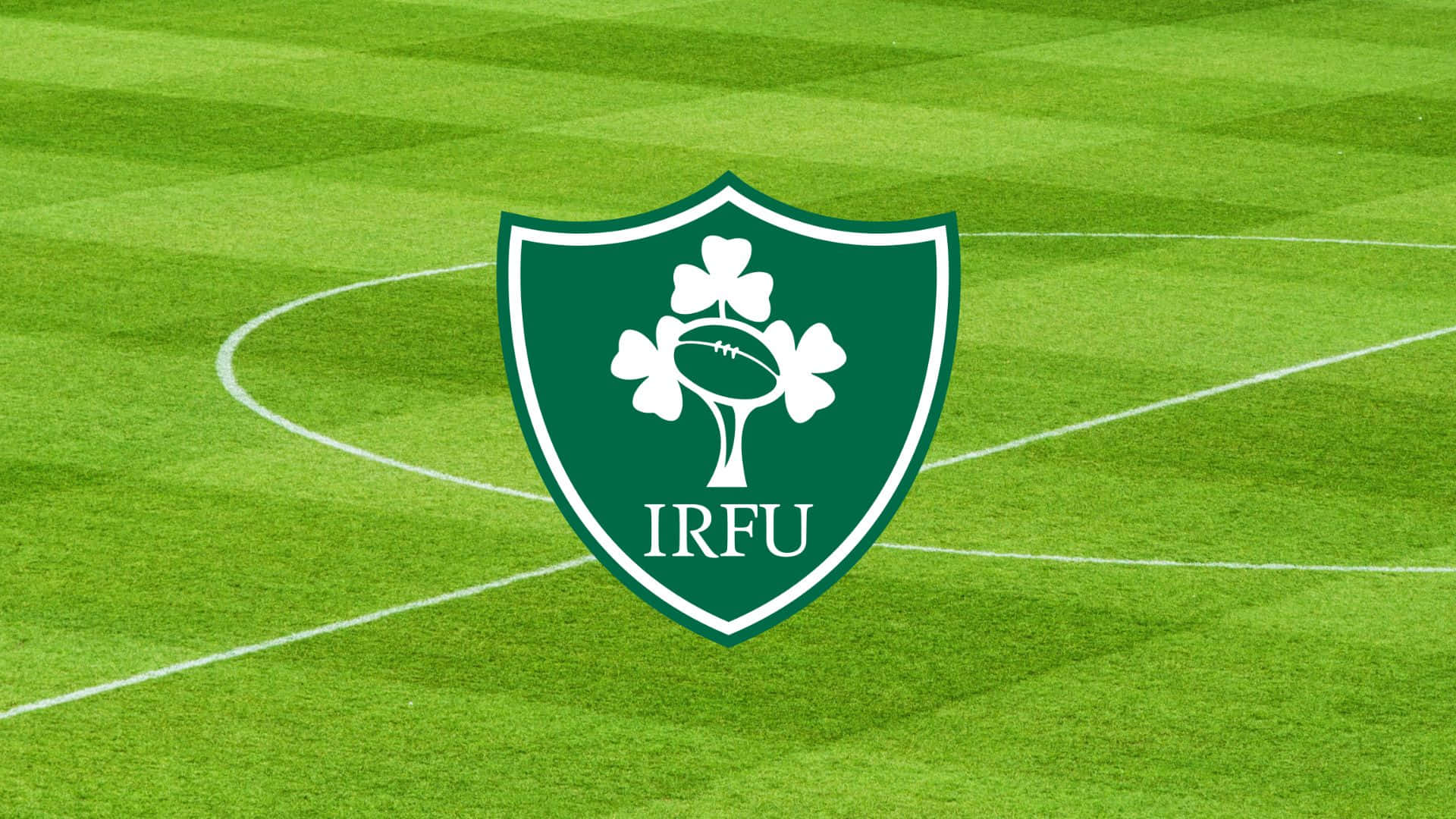 Ireland Rugby 桌布 壁紙 — 一個綠色盾牌標誌，中央有白色三葉草和橄欖球，位於充滿活力的條紋橄欖球場上。