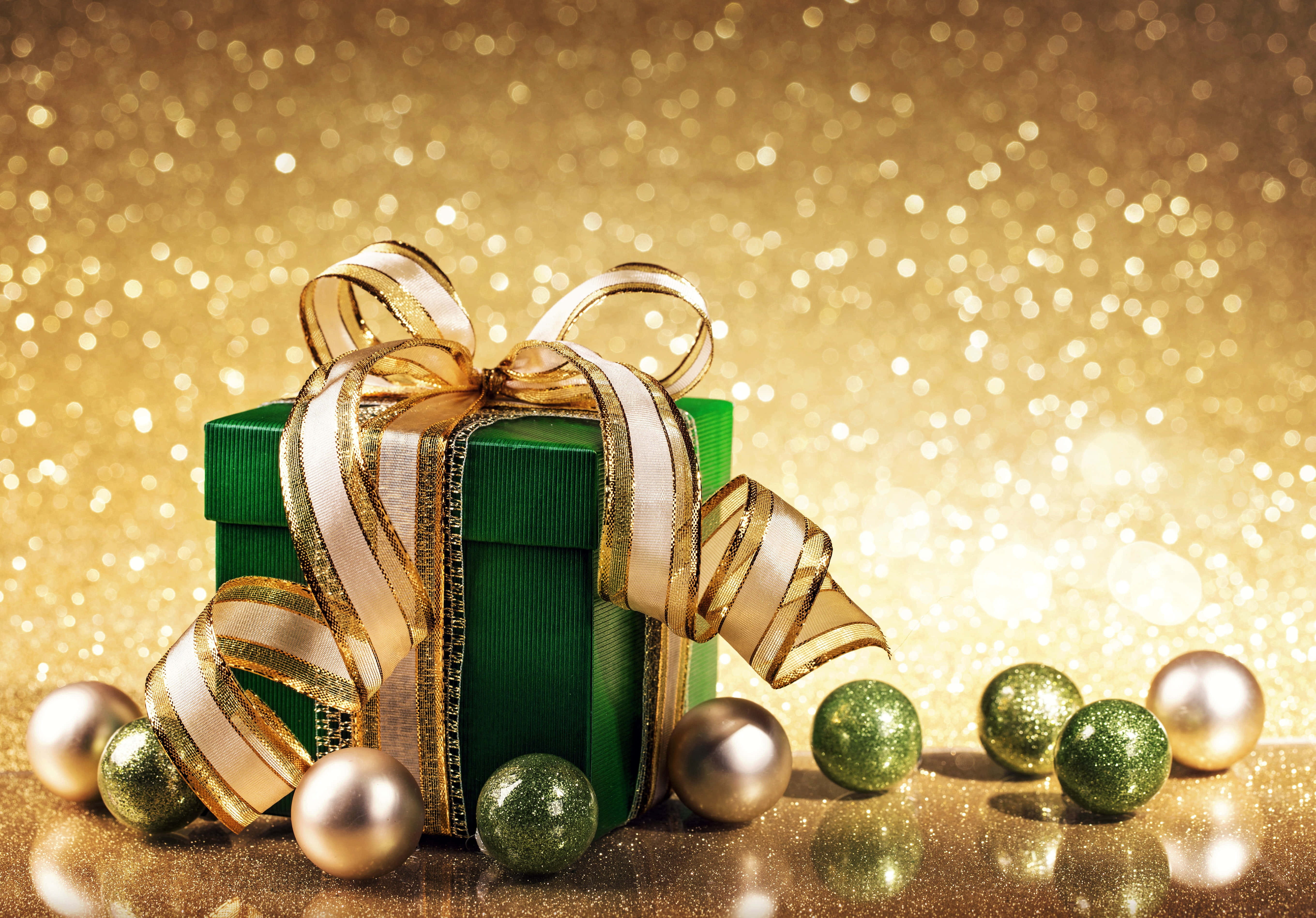 Gold Christmas 桌布 壁紙 — 一個綠色禮物盒，飾有金色和白色絲帶，周圍環繞著閃爍的綠色和金色裝飾品，背景是閃閃發光的金色。