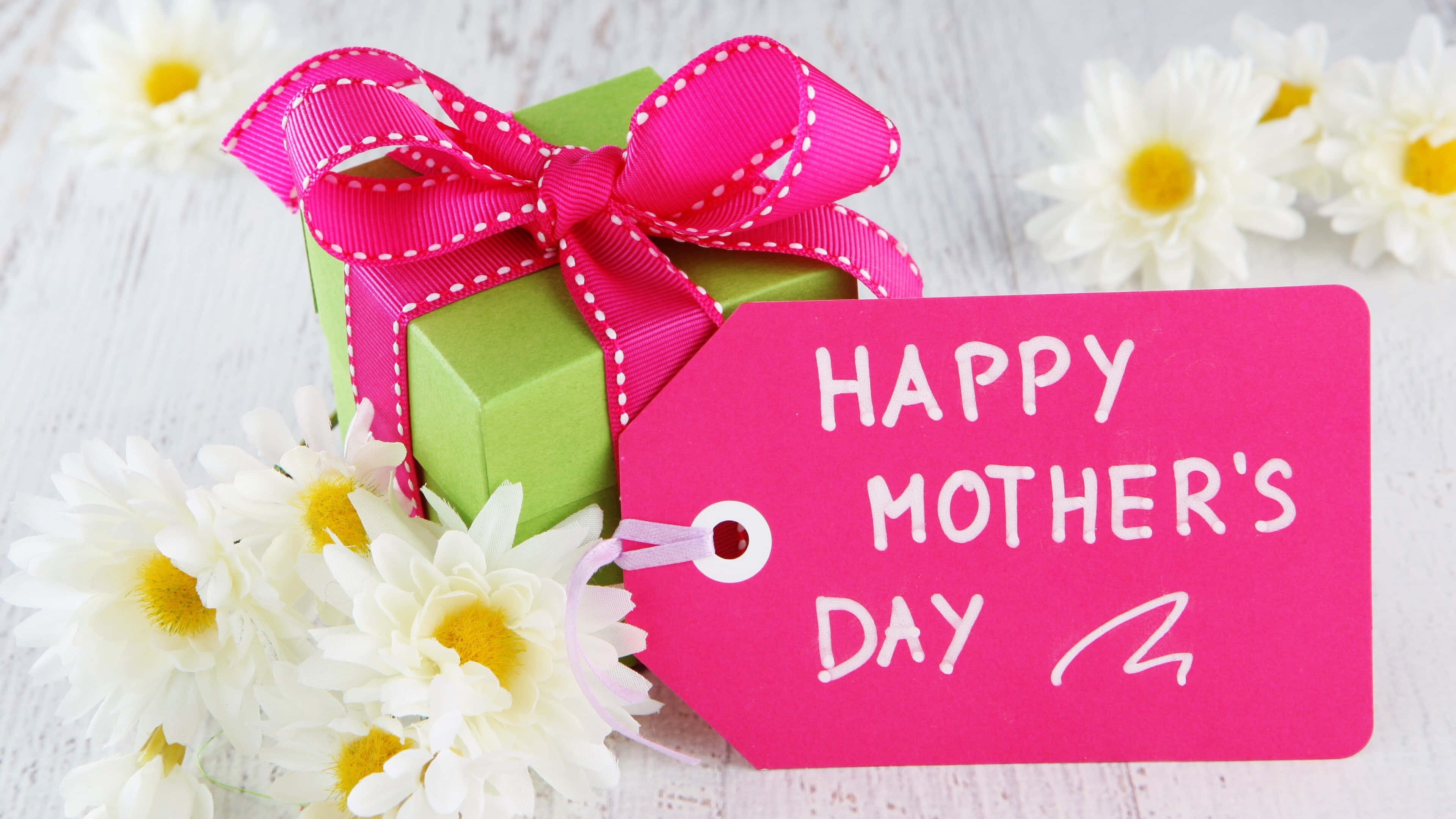 Happy Mothers Day 桌布 壁紙 — 一個綠色禮物盒綁著一條亮粉紅色絲帶，旁邊放著白色雛菊和一個寫著「Happy Mother's Day」的粉紅色標籤。