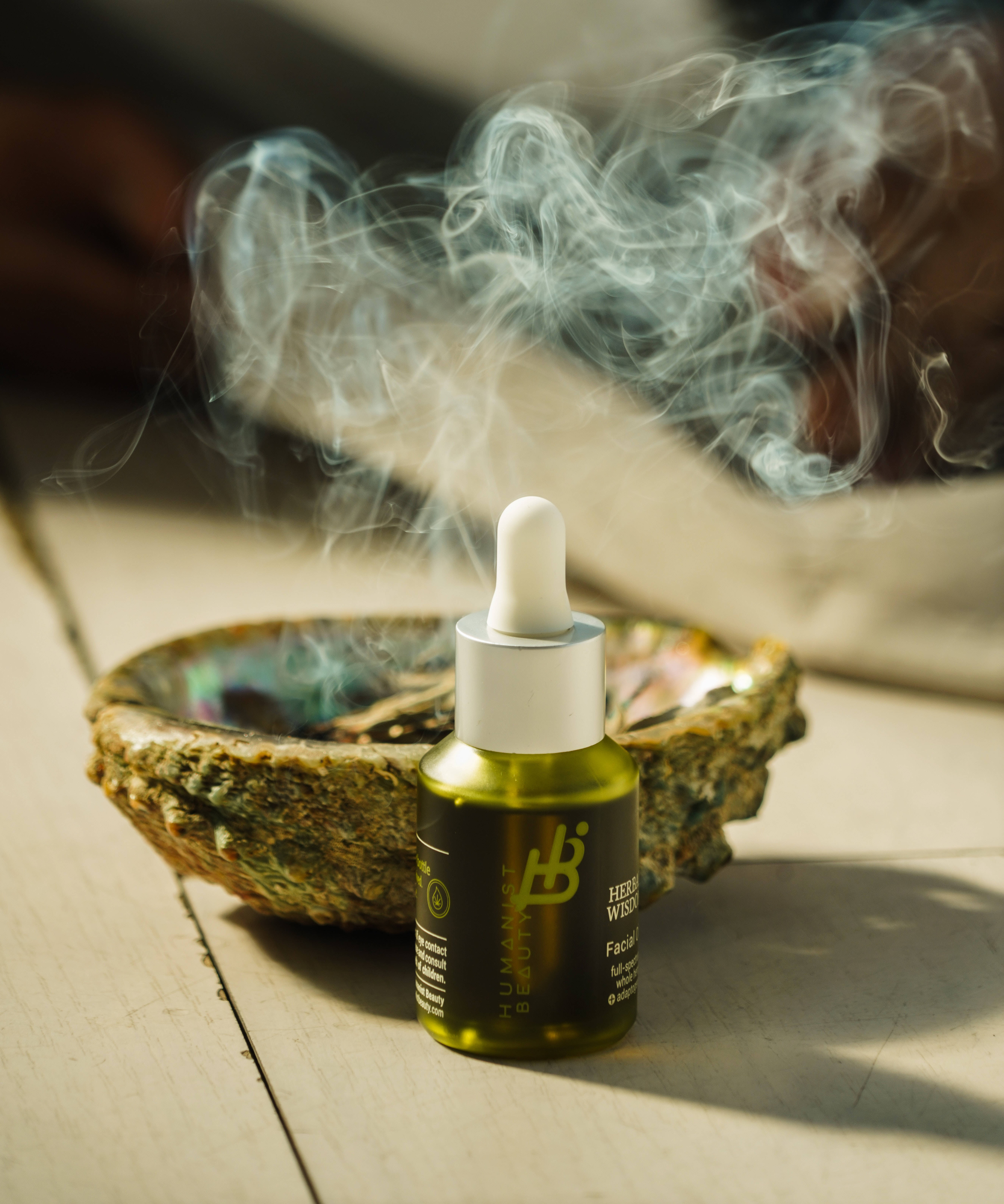 貝殼 桌布 壁紙 — 一個綠色精華瓶連滴管放在一個冒煙的鮑魚殼旁邊，木質表面沐浴在溫暖的陽光中。