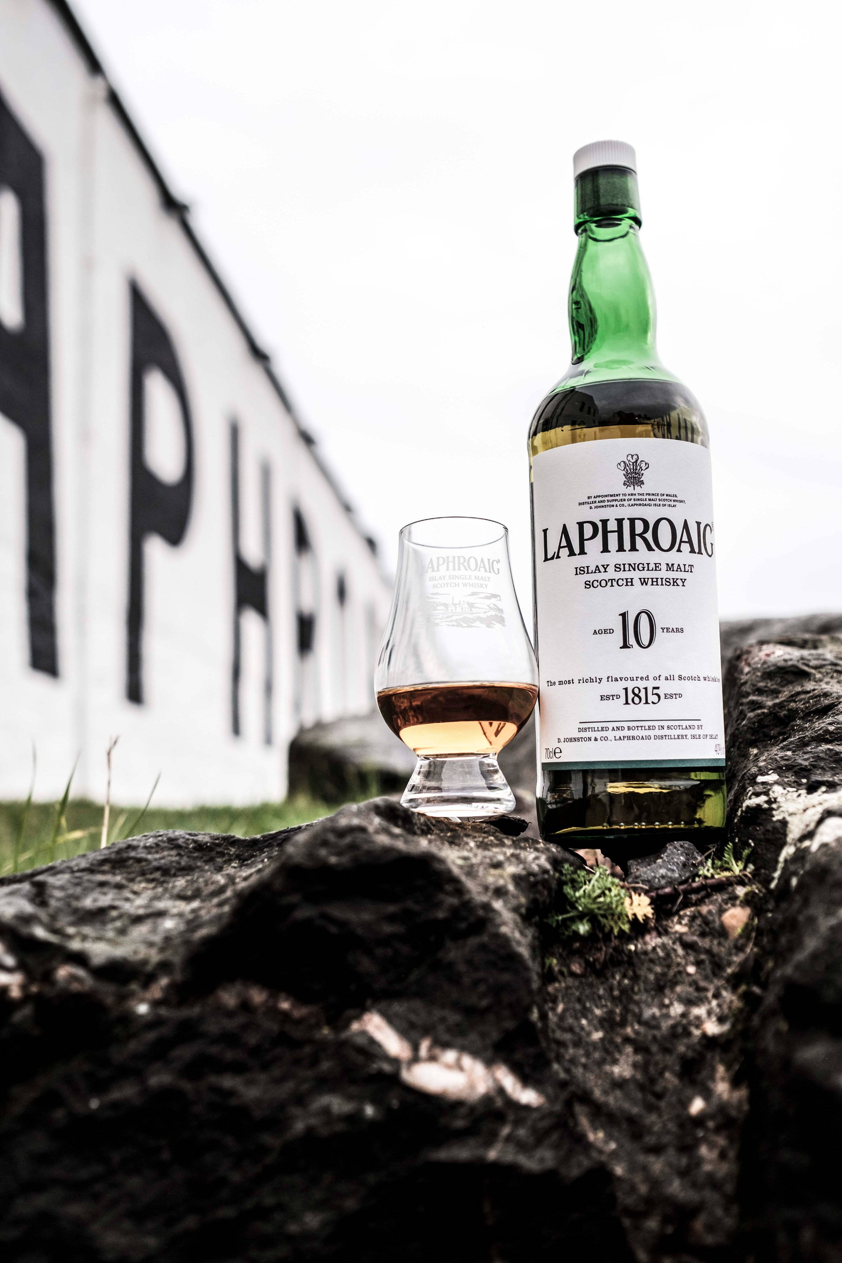 手機 桌布 壁紙 — 一個綠色酒瓶和一杯Laphroaig 10年蘇格蘭威士忌放在深色岩石上，背景是白色牆壁，上面有粗體黑色字樣。