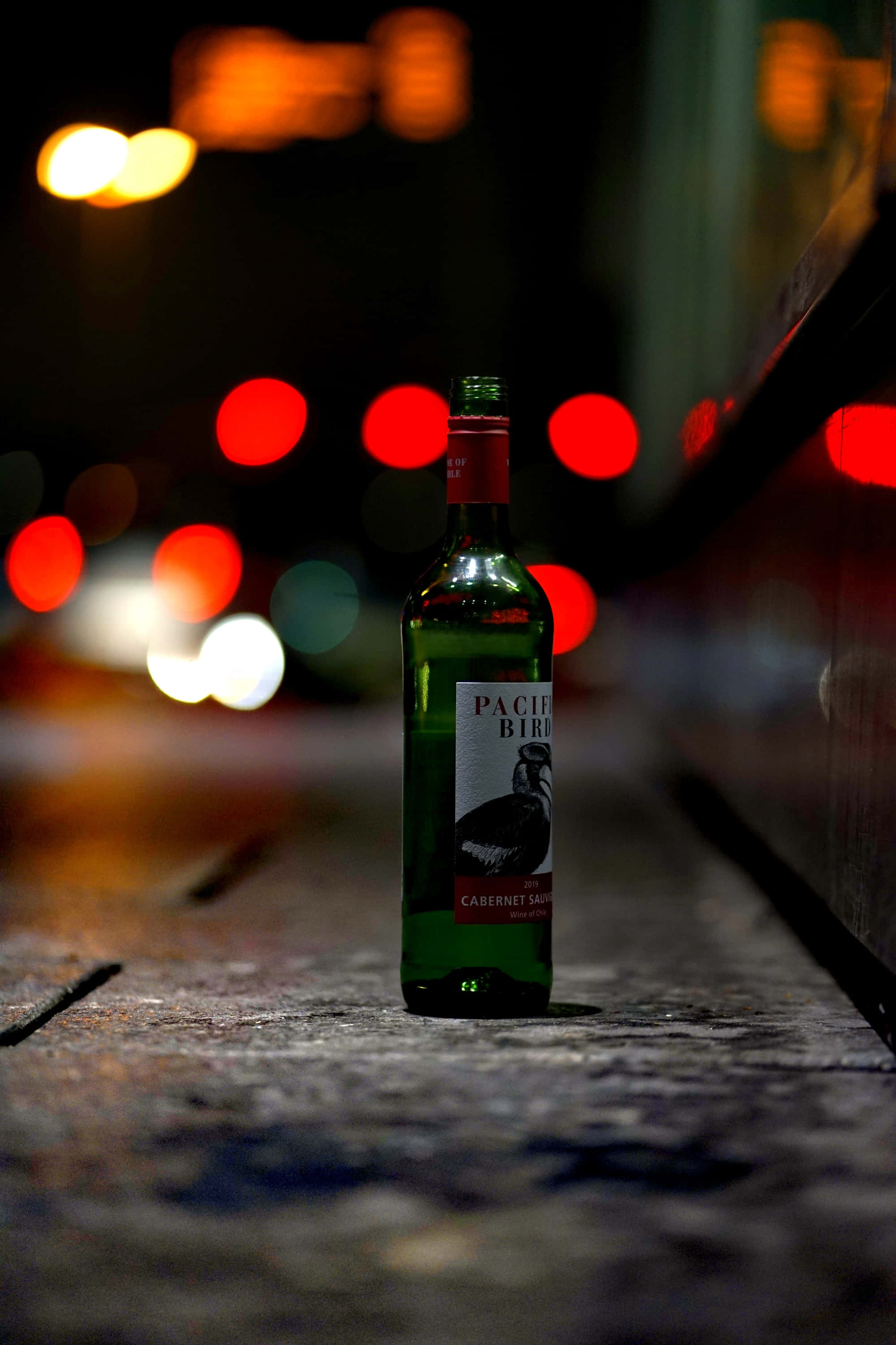 葡萄酒 桌布 壁紙 — 一個綠色酒瓶獨自立在夜晚潮濕的城市人行道上，周圍是來往車輛模糊的紅色和白色散景燈光。