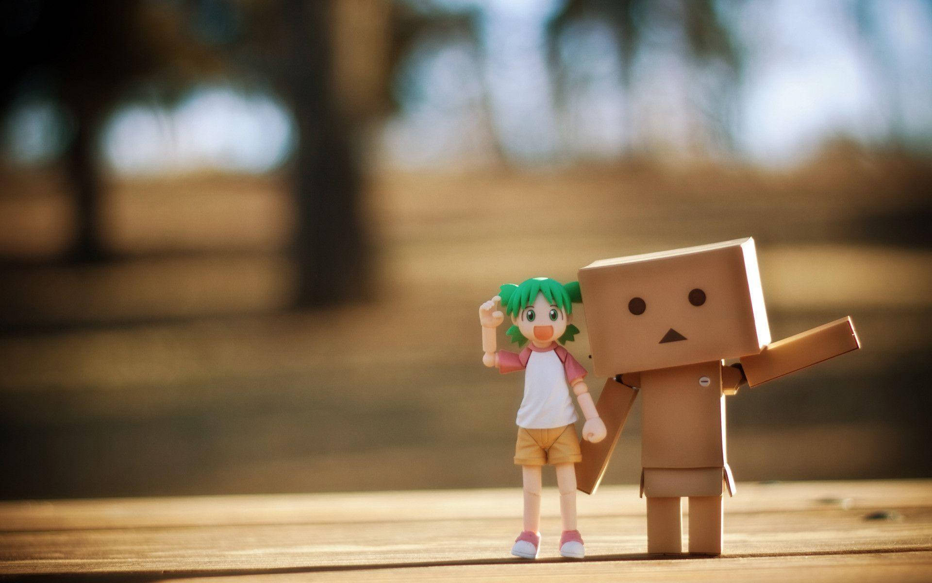 可愛的好朋友 桌布 壁紙 — 一個綠色頭髮的動漫女孩公仔站在一個紙箱機器人旁邊，放在木質表面上，背景有模糊的樹木。