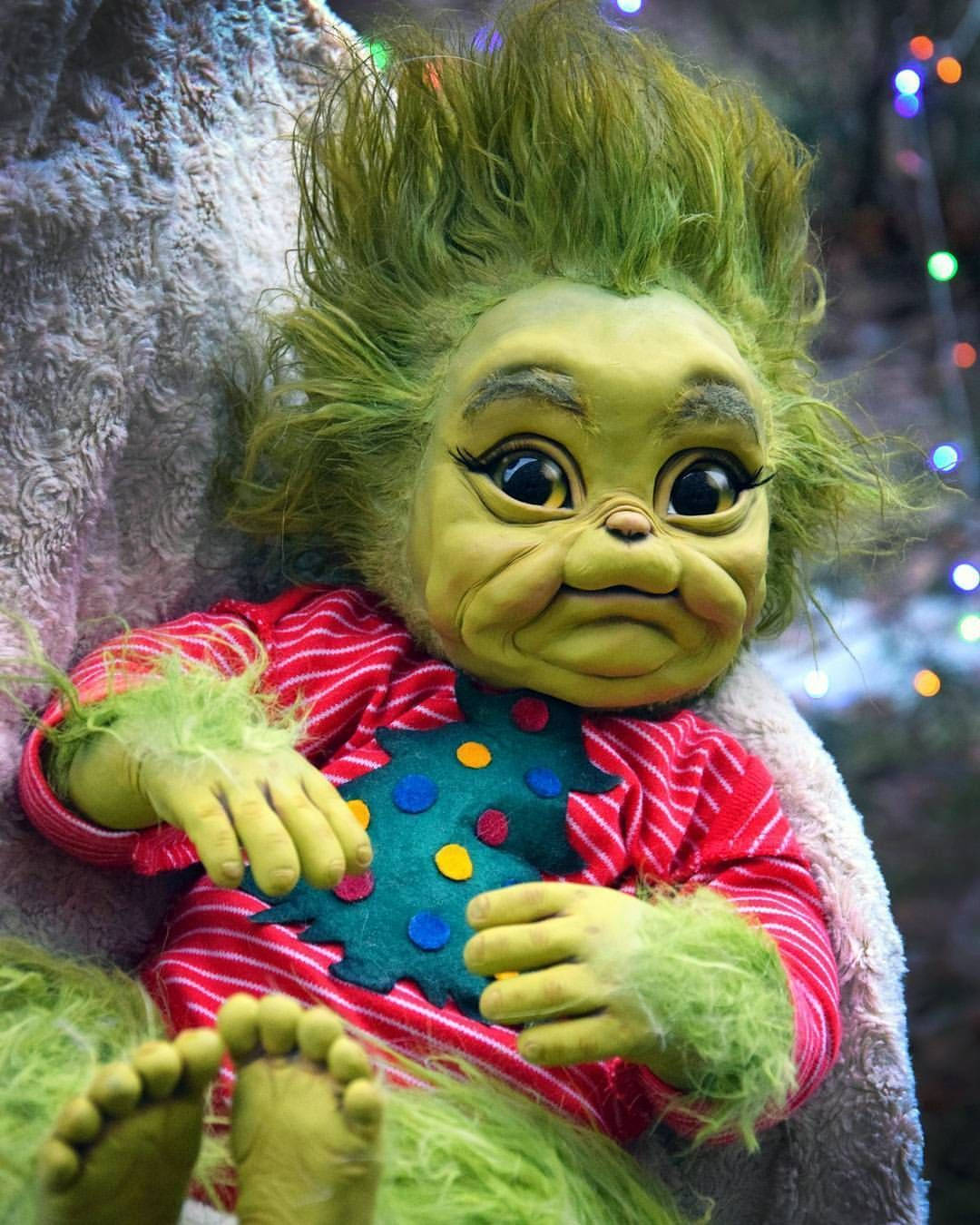 Baby Grinch 桌布 壁紙 — 一個綠色頭髮的鬼靈精公仔，穿著紅色條紋睡衣，上面有彩色圓點，坐在柔軟的白色毛毯裡，背景是閃爍的節日燈飾。