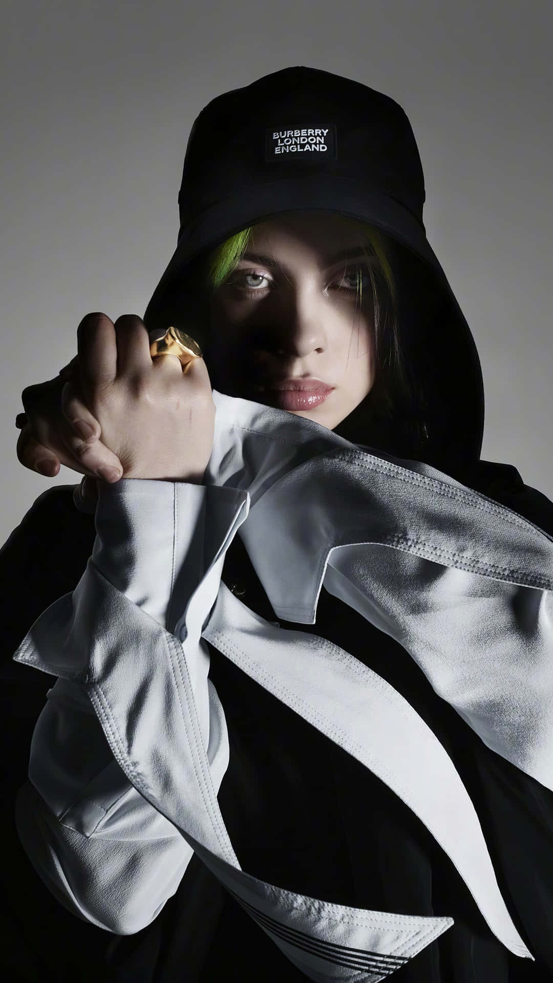 怪奇比莉 桌布 壁紙 — 一個綠髮、戴著 Burberry 漁夫帽的人，在灰色背景前緊握金戒指，眼神銳利。
