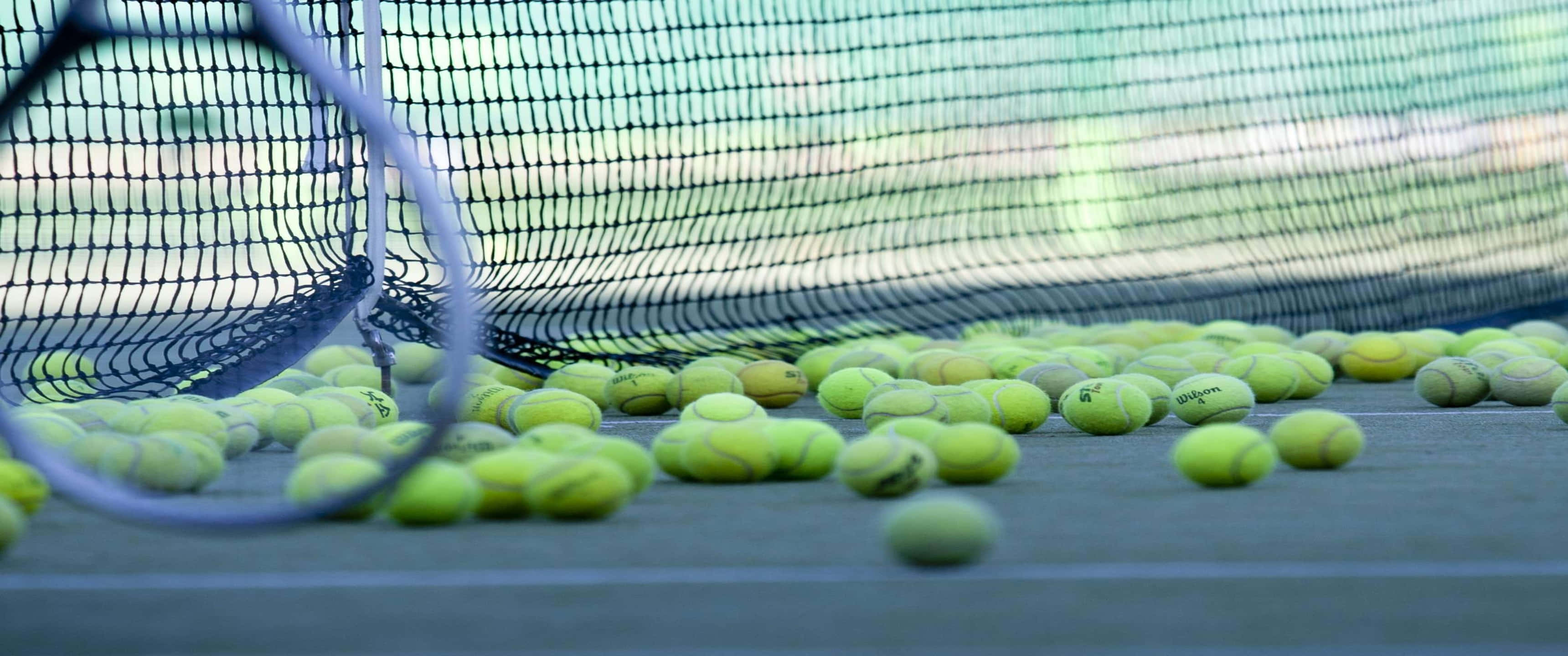 3440x1440p Tennis 桌布 壁紙 — 一個網球拍靠在網上，球場表面散落黃色網球的特寫。