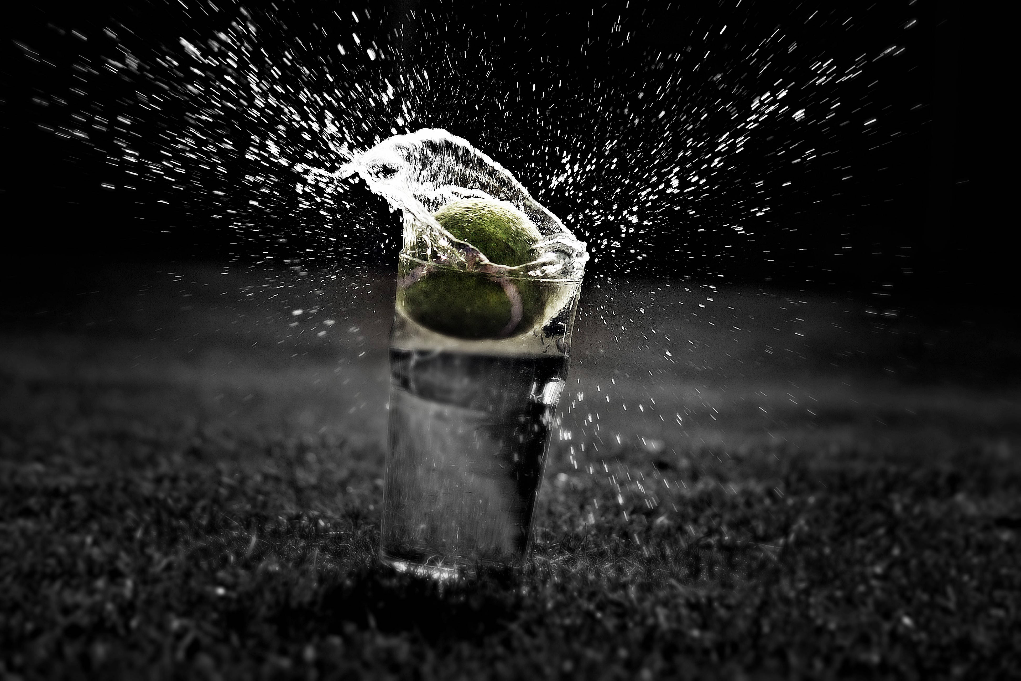 Water Splash 桌布 壁紙 — 一個網球穿過玻璃杯中的水，在深色背景上形成戲劇性的水花。