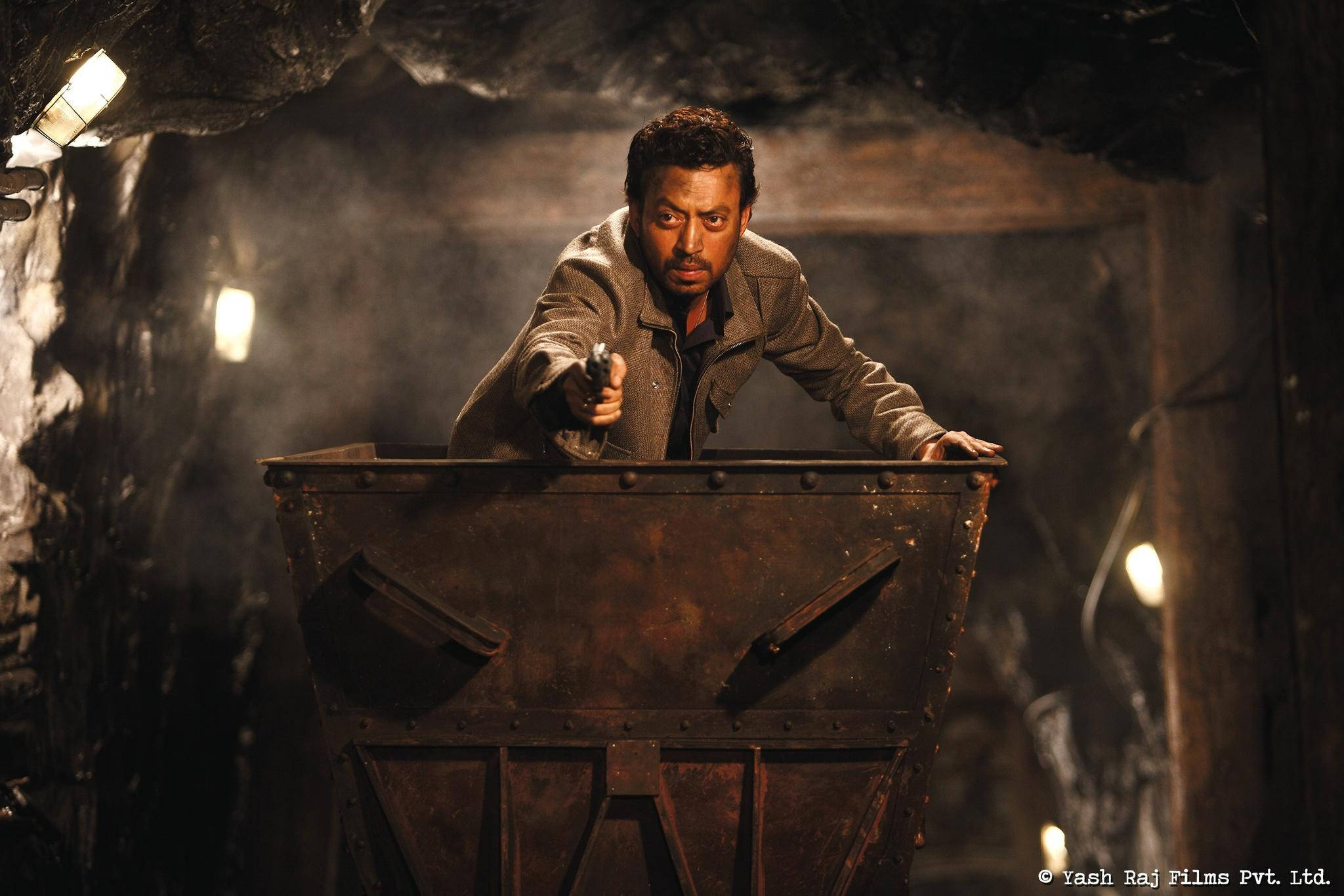 Irrfan Khan 桌布 壁紙 — 一個緊張的男人在粗糙的礦井中，從生鏽的礦車後面用一把手槍瞄準，燈光閃爍不定。