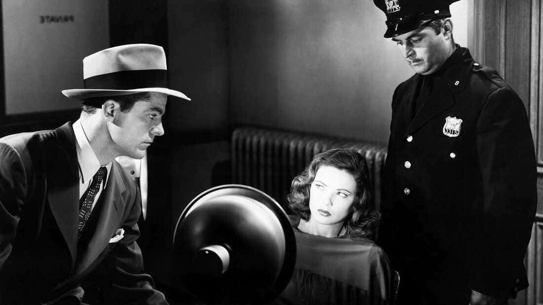 Film Noir 桌布 壁紙 — 一個緊張的黑白場景，顯示一個戴著費多拉帽的男人和一名警察站在一個坐在昏暗審訊室裏的女人上方。