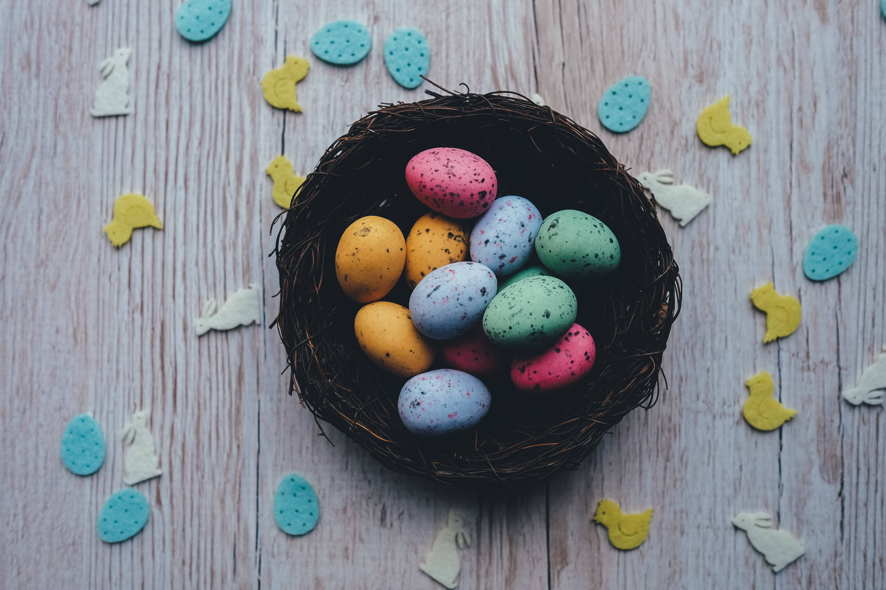 Easter 桌布 壁紙 — 一個編織的巢穴在質樸的木質表面上盛放著色彩繽紛的斑點復活節彩蛋，周圍環繞著紙製小雞和雞蛋。