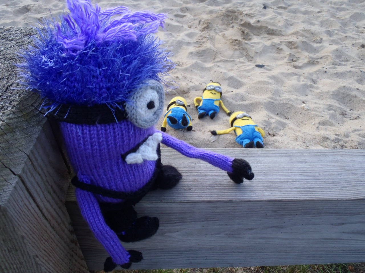 evil 桌布 壁紙 — 一個編織的紫色Minion，頭髮凌亂，靠在木欄杆上，而三個黃色Minion在後面的沙灘上睡覺。