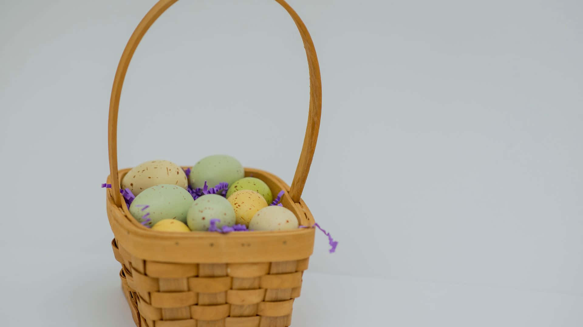 Easter Basket 桌布 壁紙 — 一個編織籃子，裡面裝滿了粉彩斑點蛋，放在紫色碎紙上，背景是純白色。
