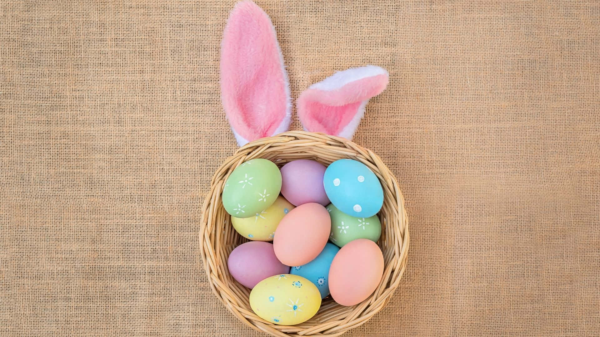 Easter Bunny 桌布 壁紙 — 一個編織籃子帶有粉紅色兔耳，在質感粗麻布背景上盛放著柔和色彩的復活節蛋。
