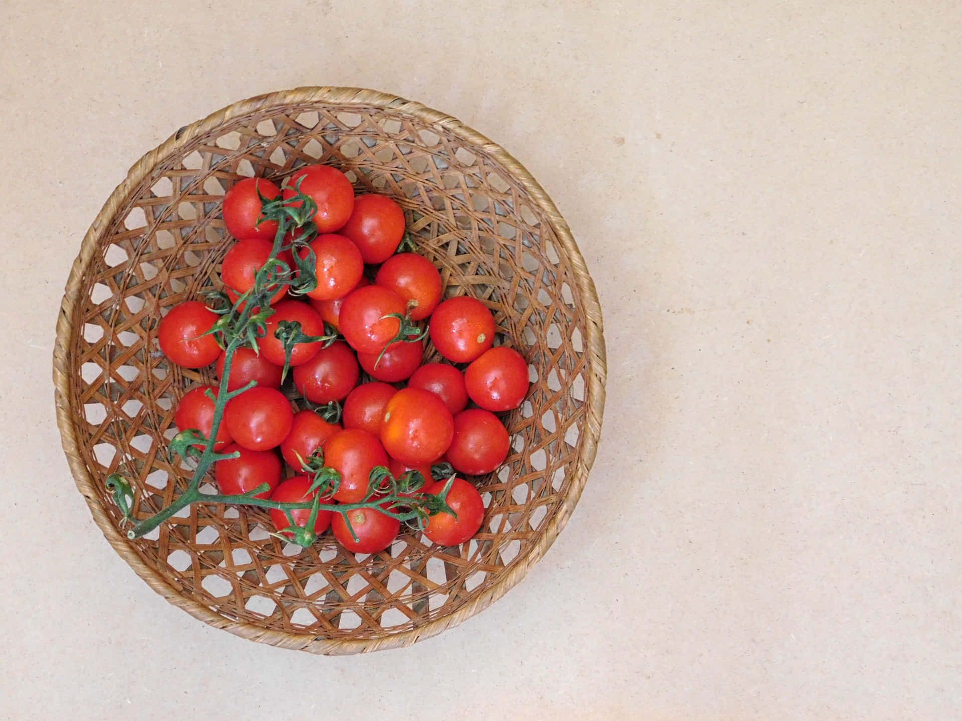 Tomatoes 壁紙 — 一個編織籃子盛著鮮紅色的櫻桃番茄，仍然連著綠色的藤蔓，放在淺米色的表面上。