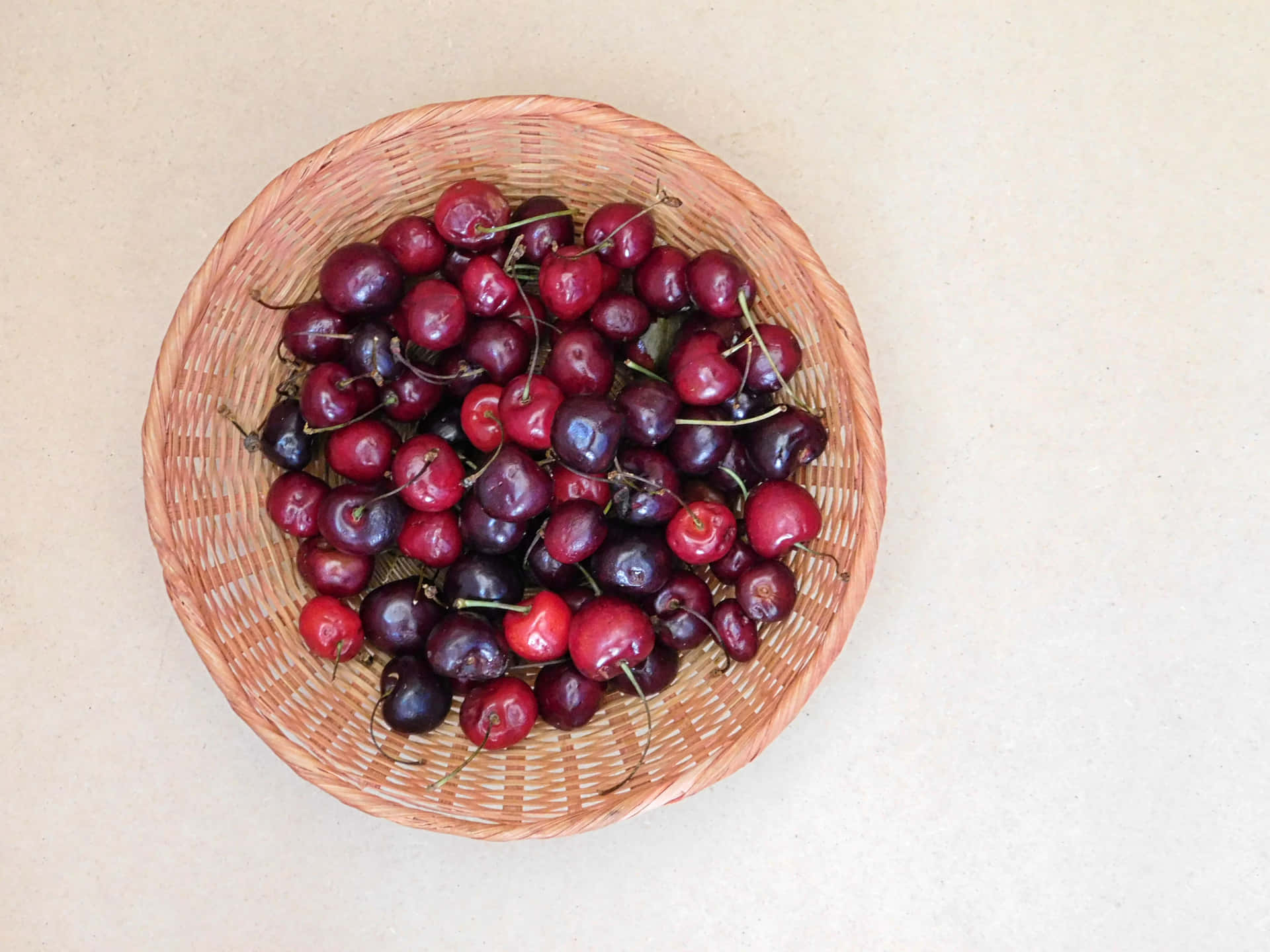 Cherries 壁紙 — 一個編織籃子裝滿了飽滿的深紅色櫻桃，帶有莖，背景為中性米色。