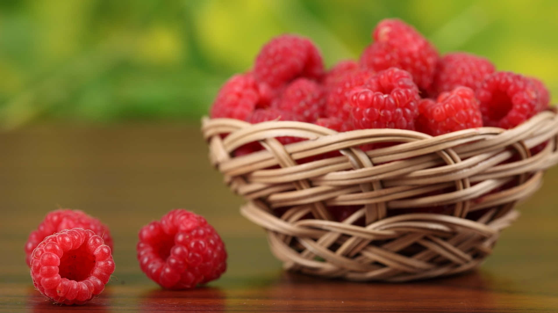 Raspberry 桌布 壁紙 — 一個編織籃子裝滿了飽滿的紅覆盆子，放在木桌上，旁邊有兩顆莓果，背景是柔和的綠色。