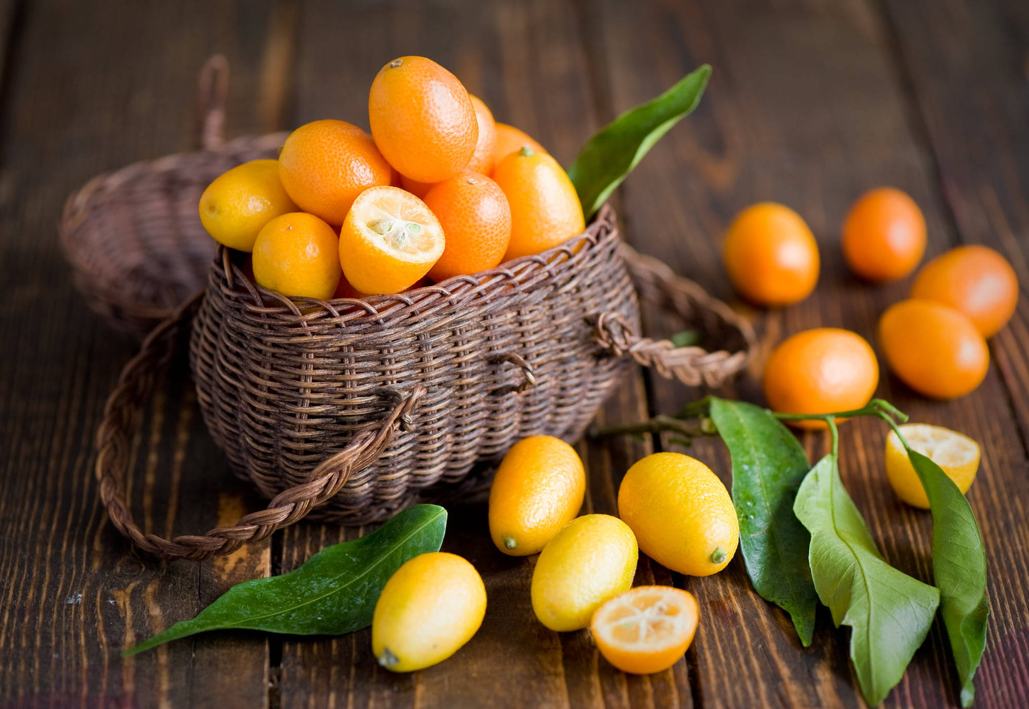 Kumquat 桌布 壁紙 — 一個編織籃子裝滿了鮮豔的橙色金橘和新鮮的綠葉，放在質樸的木桌上，散落著水果和切開的半邊，露出多汁的果肉。