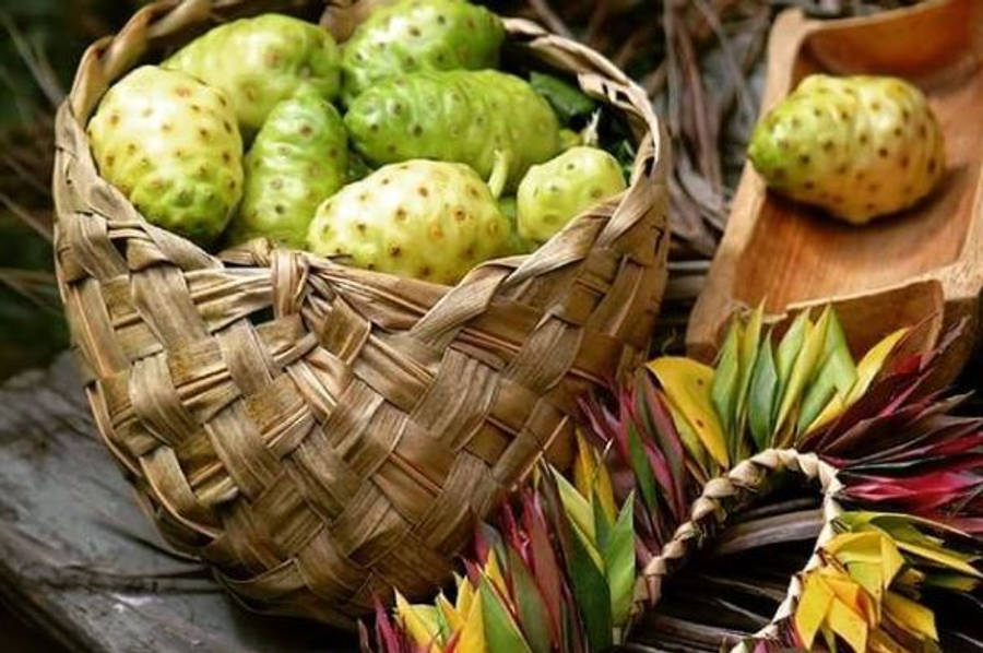 Fruits 桌布 壁紙 — 一個編織籃子裝滿帶刺的綠色諾麗果，旁邊有木托盤和色彩繽紛的熱帶葉子。