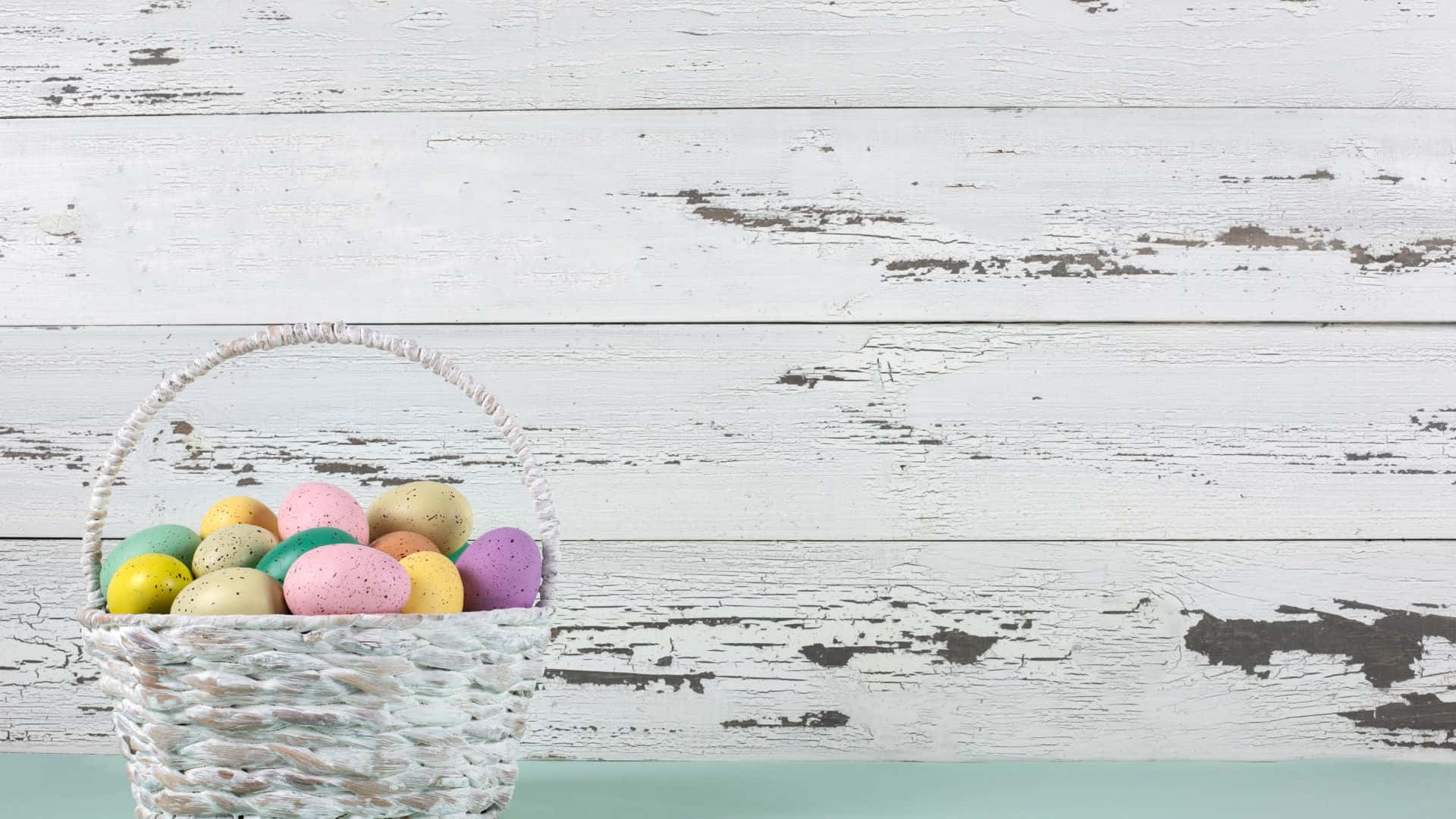 Easter Basket 桌布 壁紙 — 一個編織籃子裝滿色彩繽紛的斑點復活節蛋，放在風化的白色木質表面上。