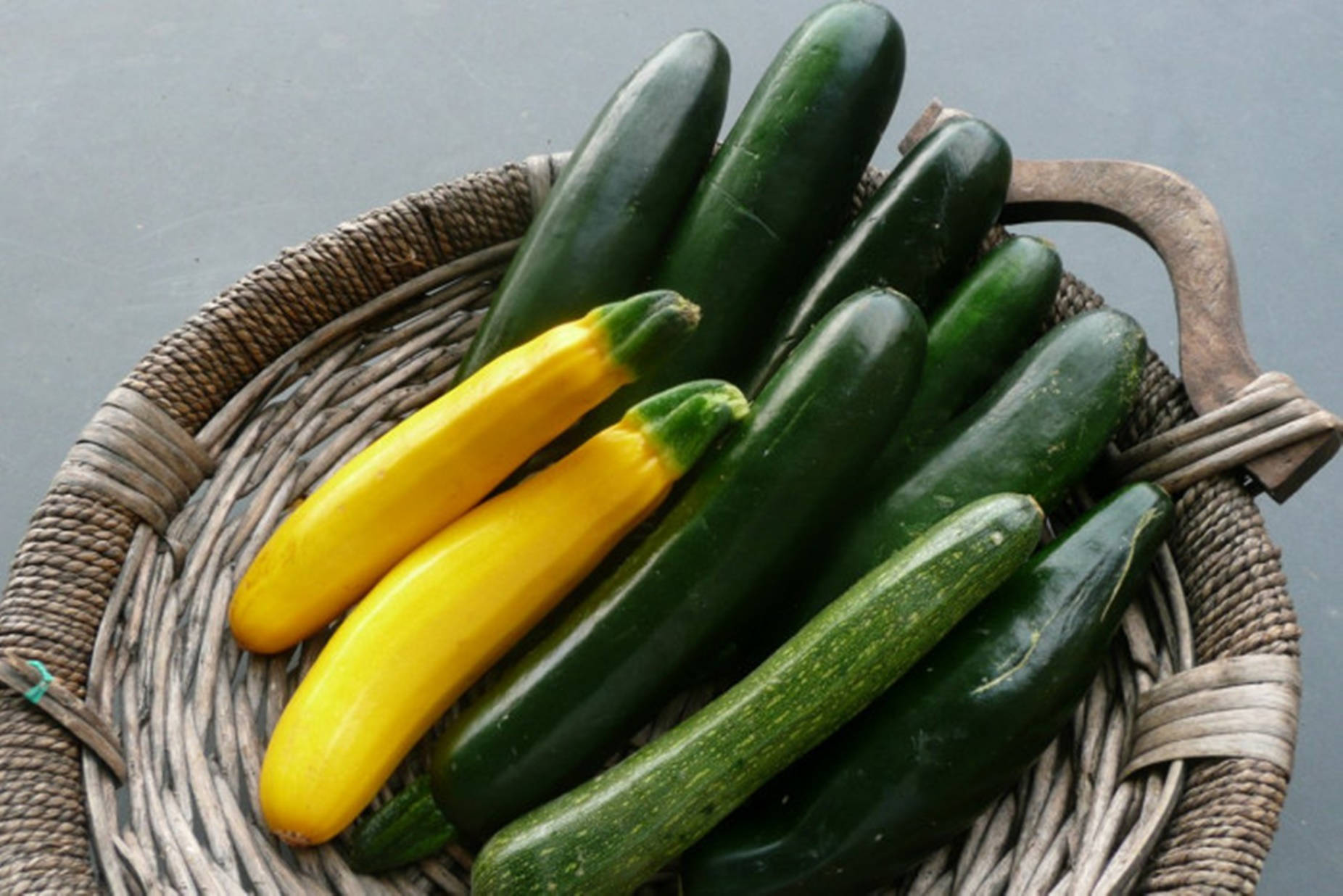 Zucchini 桌布 壁紙 — 一個編織籃子裝著新鮮的綠色櫛瓜和鮮黃色的夏季南瓜，背景為柔和的灰色。