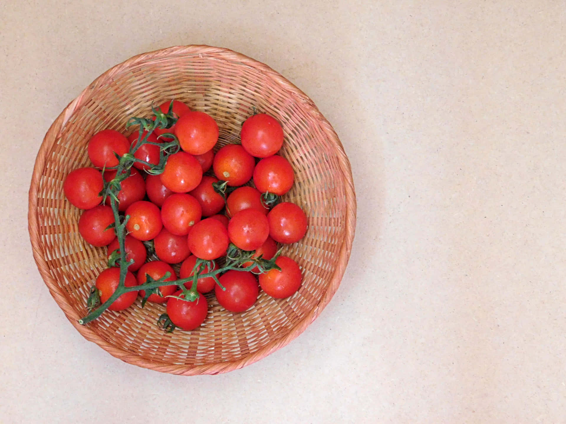 Tomatoes 壁紙 — 一個編織籃子裝著鮮紅色的櫻桃番茄，仍然連著綠色的藤蔓，放在淺米色的表面上。