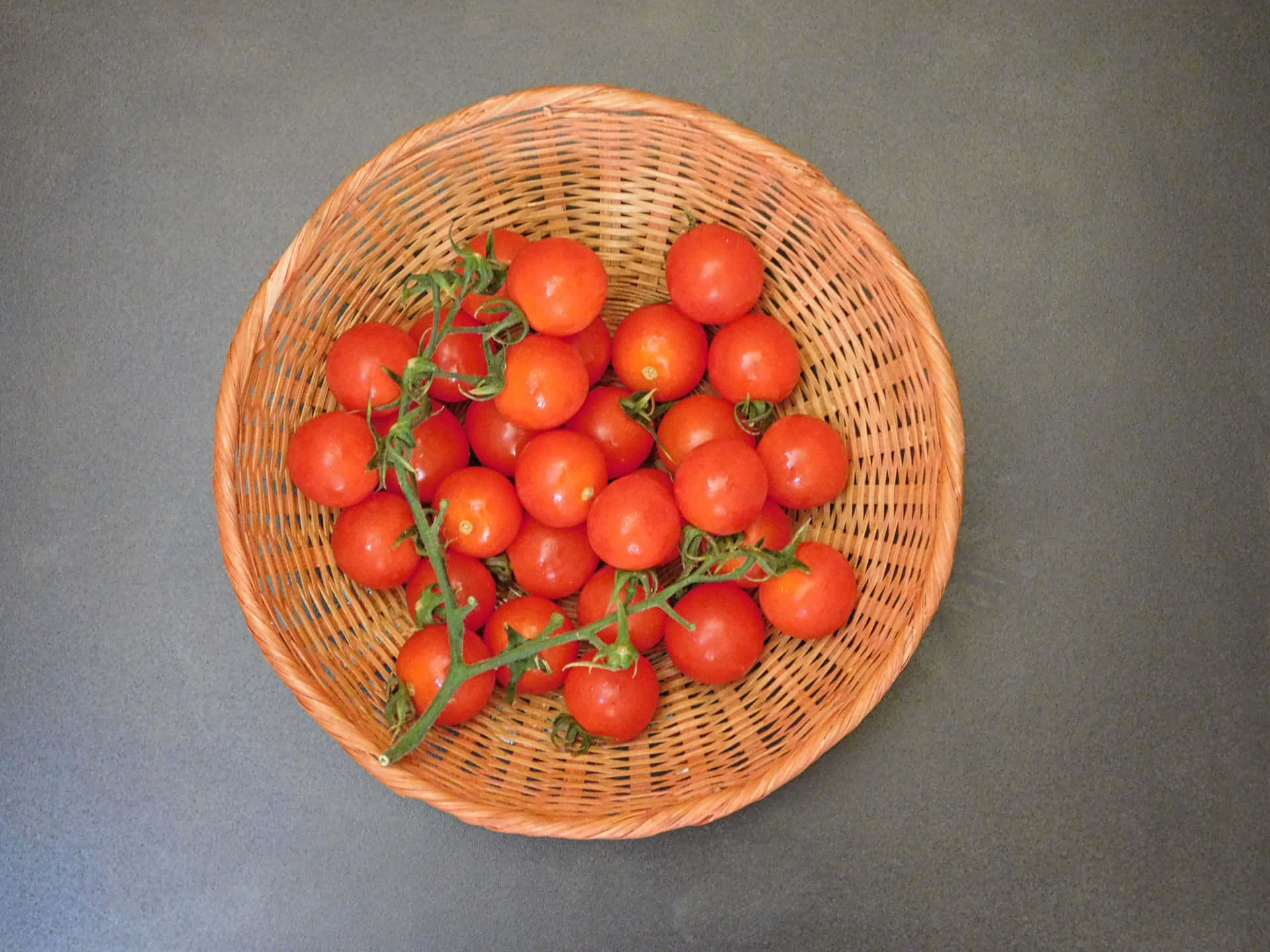 Tomatoes 壁紙 — 一個編織籃子裝著鮮紅色的櫻桃番茄，仍然連著綠色的藤蔓，放在柔和的灰色背景前。