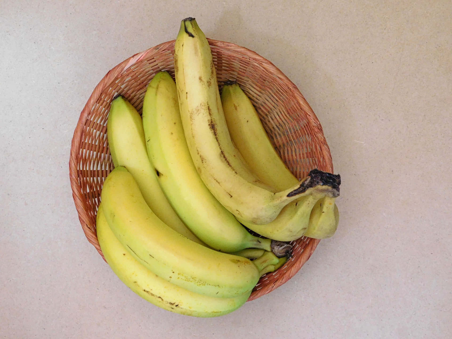 Bananas 壁紙 — 一個編織籃子裡放著一串成熟的黃色香蕉，上面有棕色斑點，放在淺米色檯面上。