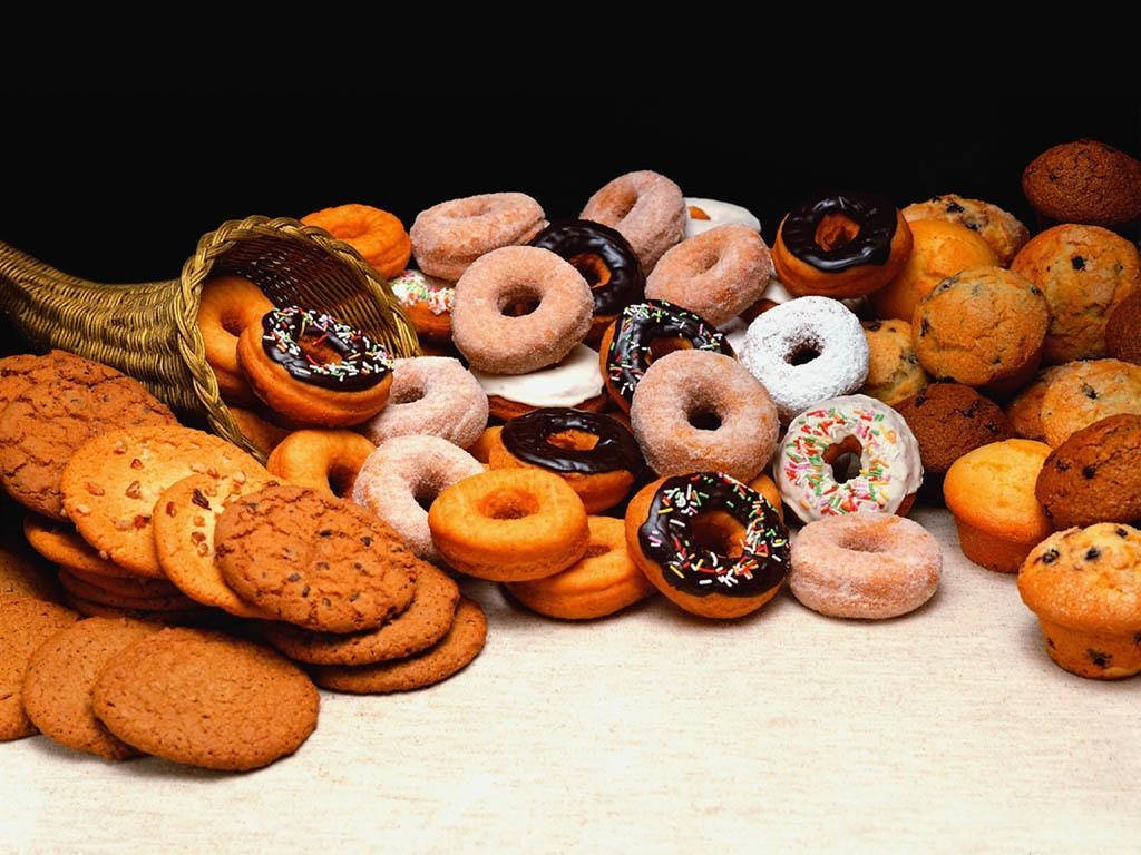 Donut 桌布 壁紙 — 一個編織籃子裡溢出各種甜甜圈、鬆餅和餅乾，散落在淺色木質表面上，場面豐盛。