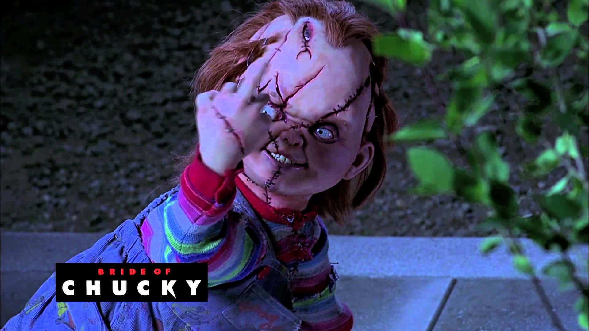 Chucky Doll 桌布 壁紙 — 一個縫合的公仔，紅色頭髮，帶著邪惡的笑容，在黑暗的戶外環境中舉起一隻血淋淋的手。