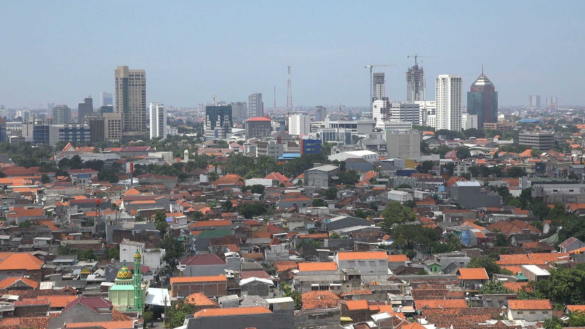 Jakarta 桌布 壁紙 — 一個繁忙城市景觀的全景視圖，融合了傳統紅瓦屋頂住宅與現代高樓大廈，在清澈的藍天下。