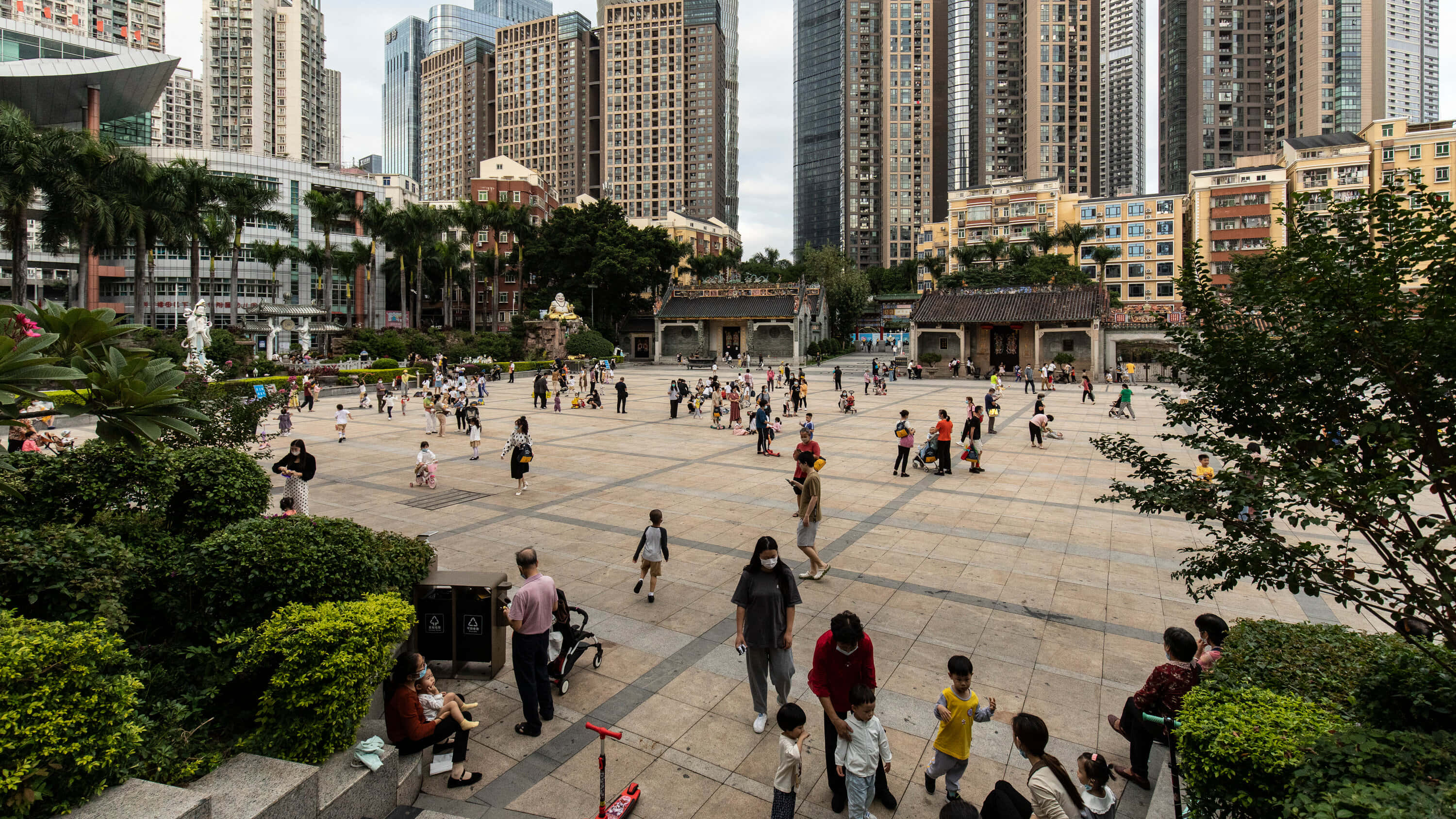 中國 桌布 壁紙 — 一個繁忙的城市廣場，人們在陰天之下，於綠化和高聳的摩天大樓之間漫步和玩耍。