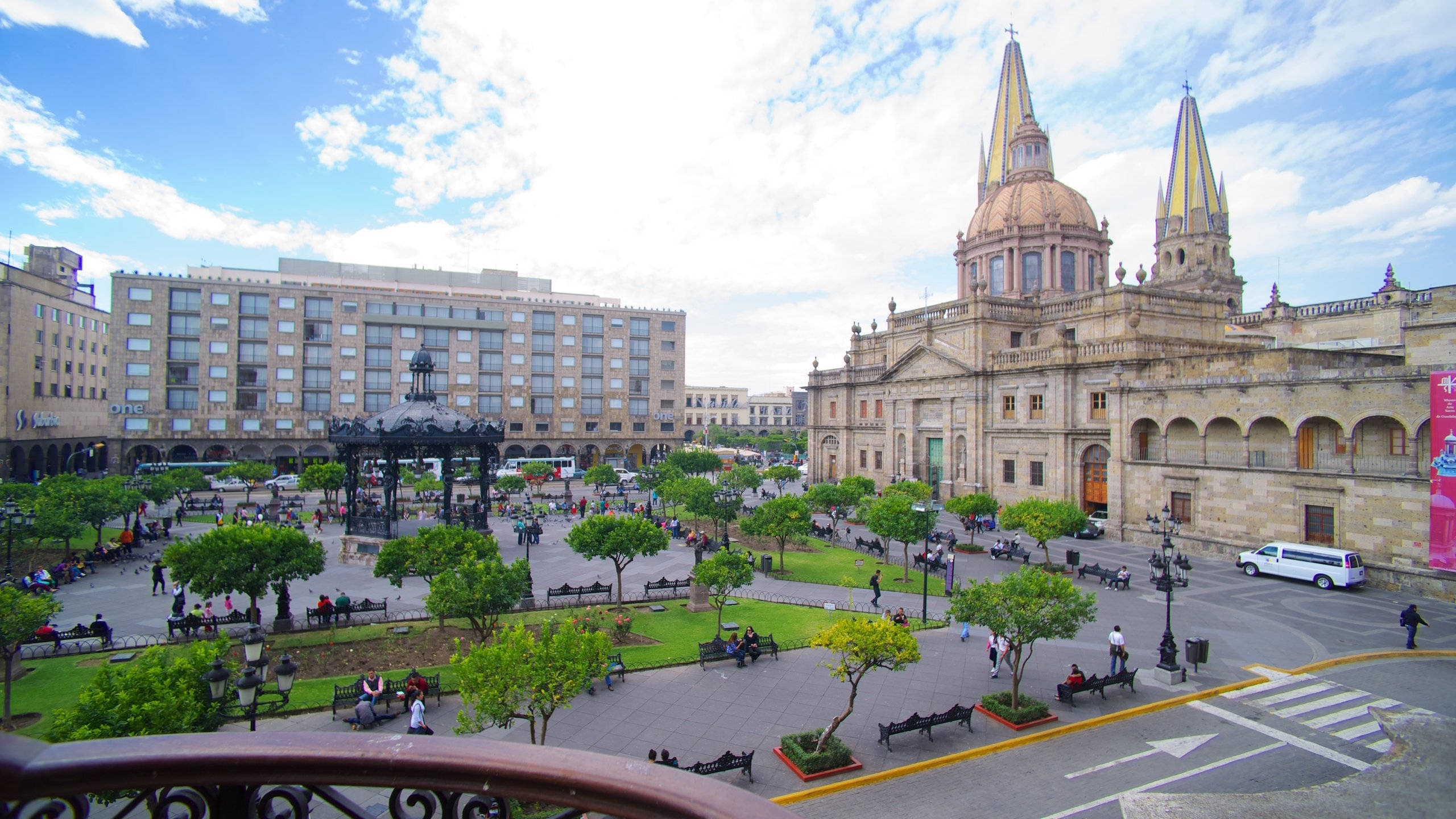 Guadalajara 桌布 壁紙 — 一個繁忙的城市廣場，有一座帶有雙塔的大教堂和一個中央噴泉，周圍環繞著綠樹，人們在部分多雲的天空下在長椅上放鬆。