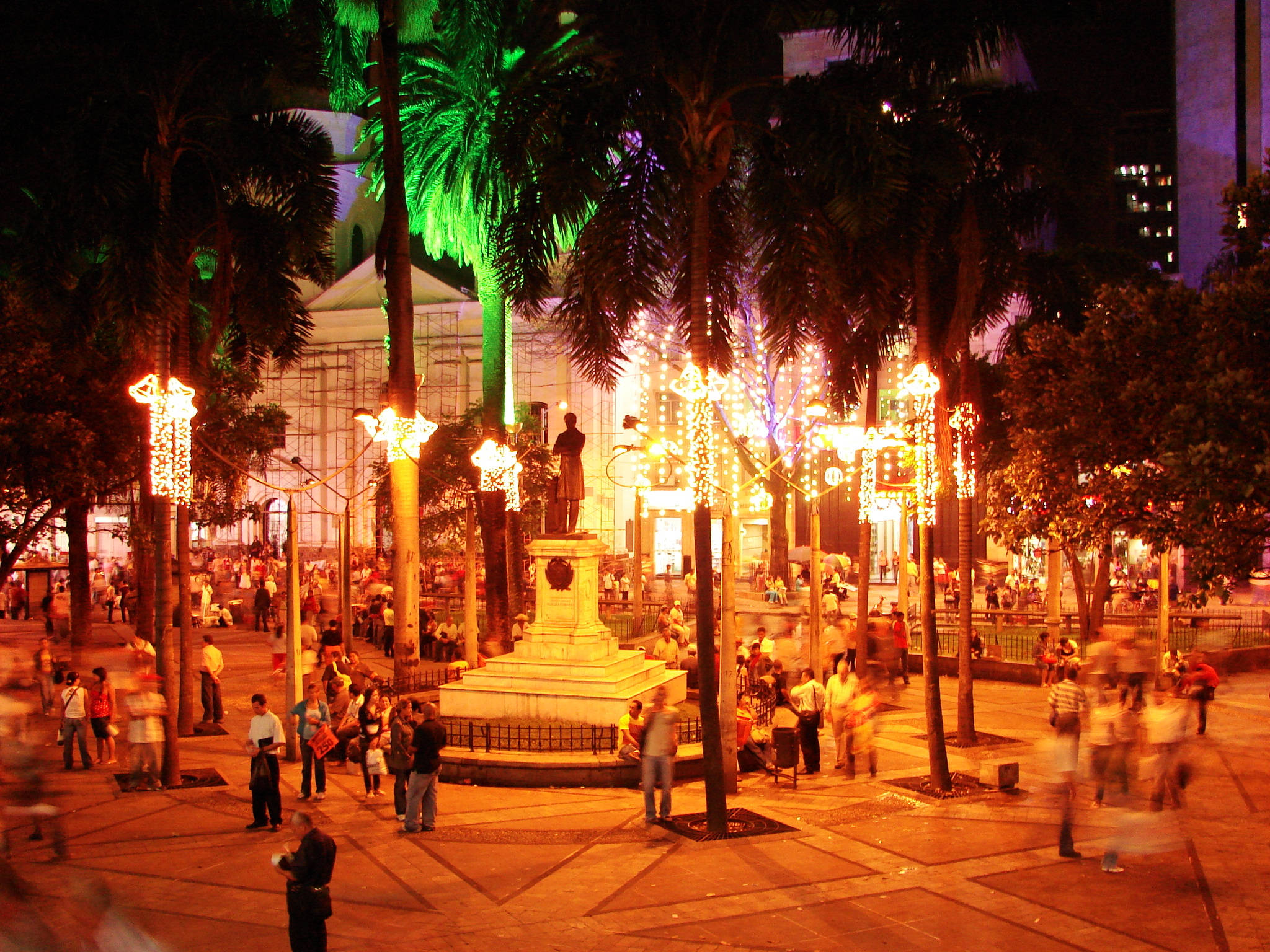 Medellin 桌布 壁紙 — 一個繁忙的城市廣場在夜晚被溫暖的串燈和發光的棕櫚樹照亮，人們在中央紀念碑周圍漫步。