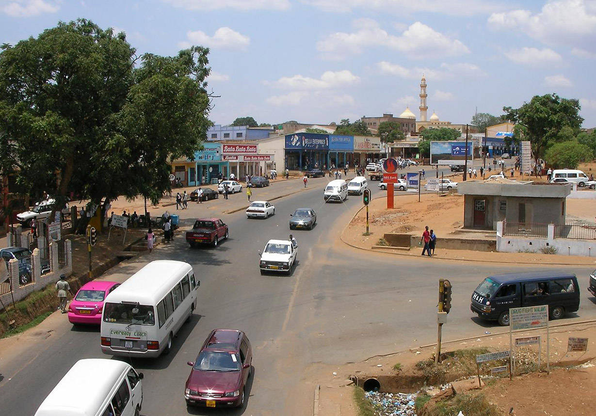Malawi 桌布 壁紙 — 一個繁忙的城市街道交叉口，有汽車、行人和色彩繽紛的建築物，天空明亮且部分多雲。