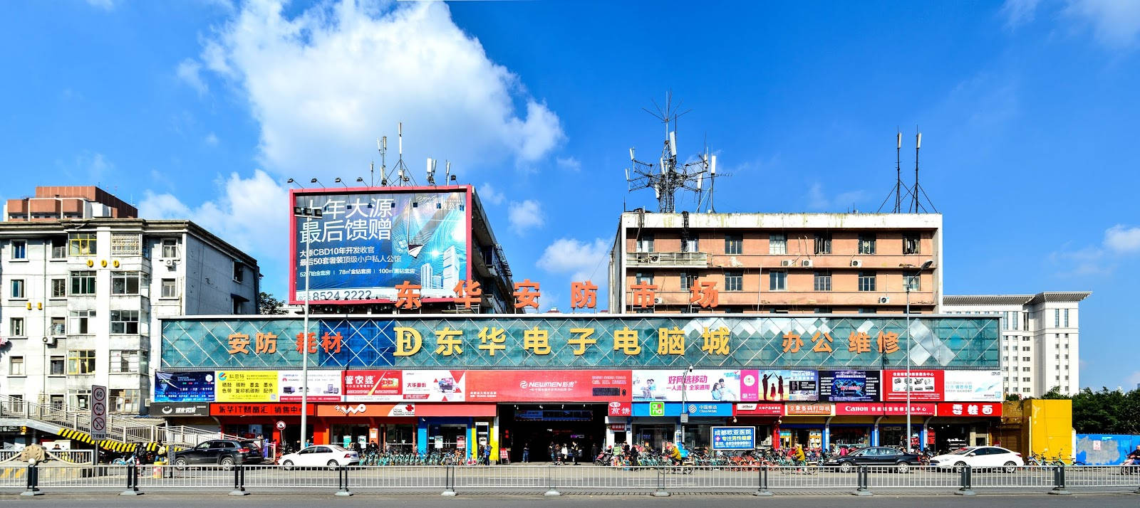Chengdu 桌布 壁紙 — 一個繁忙的城市電子市場，在明亮的藍天下，有散落的雲朵和裝飾著彩色招牌和天線的高聳建築物。