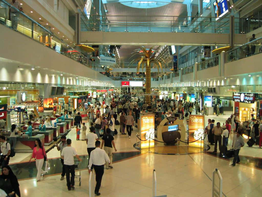 Shopping 桌布 壁紙 — 一個繁忙的多層機場航廈，旅客在明亮的燈光下經過商店和資訊亭。
