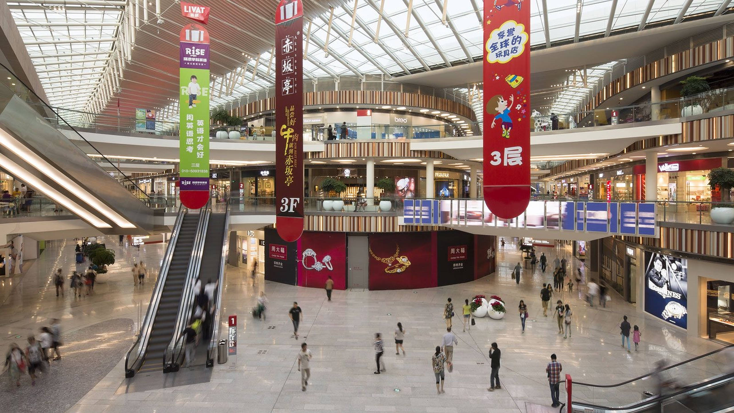 Mall 桌布 壁紙 — 一個繁忙的多層購物商場，設有自動扶梯、購物者和色彩繽紛的橫幅，上方是玻璃天花板。