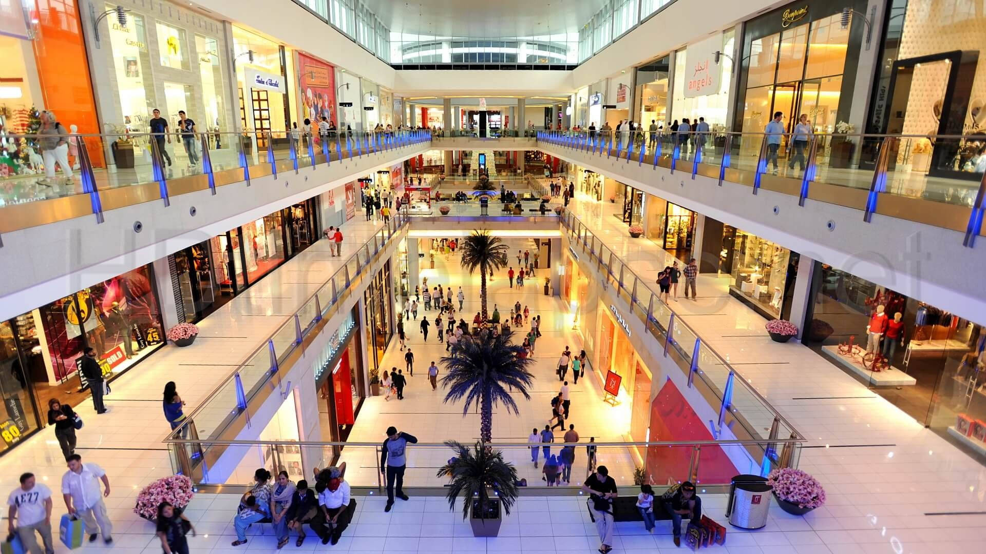 Mall 桌布 壁紙 — 一個繁忙的多層購物商場，購物者在中庭的商店和棕櫚樹之間走動。