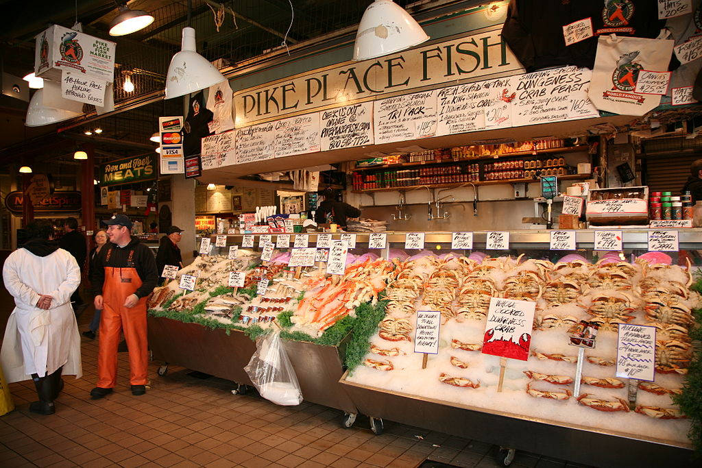 Pike Place Market 桌布 壁紙 — 一個繁忙的室內魚市場攤位展示冰上新鮮海鮮，附有手寫標牌，身穿圍裙的工人正在接待顧客。