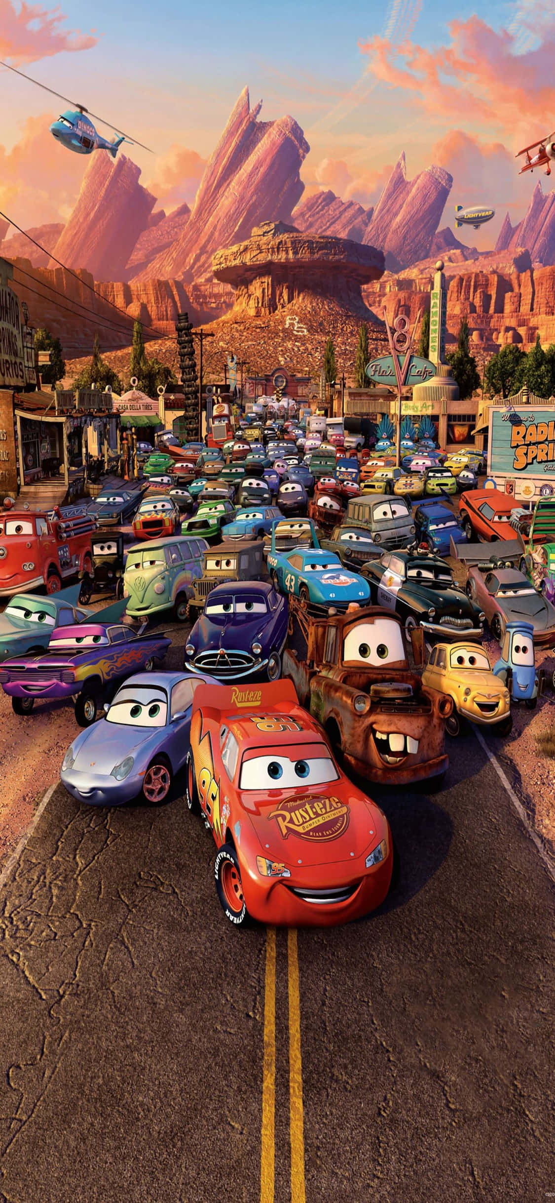 Lightning Mcqueen 桌布 壁紙 — 一個繁忙的沙漠小鎮，充滿擬人化的汽車，背景是溫暖的日落天空和岩石山脈。