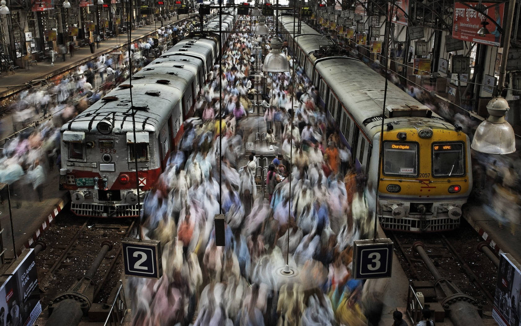 Mumbai 桌布 壁紙 — 一個繁忙的火車站月台，擠滿了模糊的人群，在懸掛的燈光下在兩列靜止的火車之間移動。
