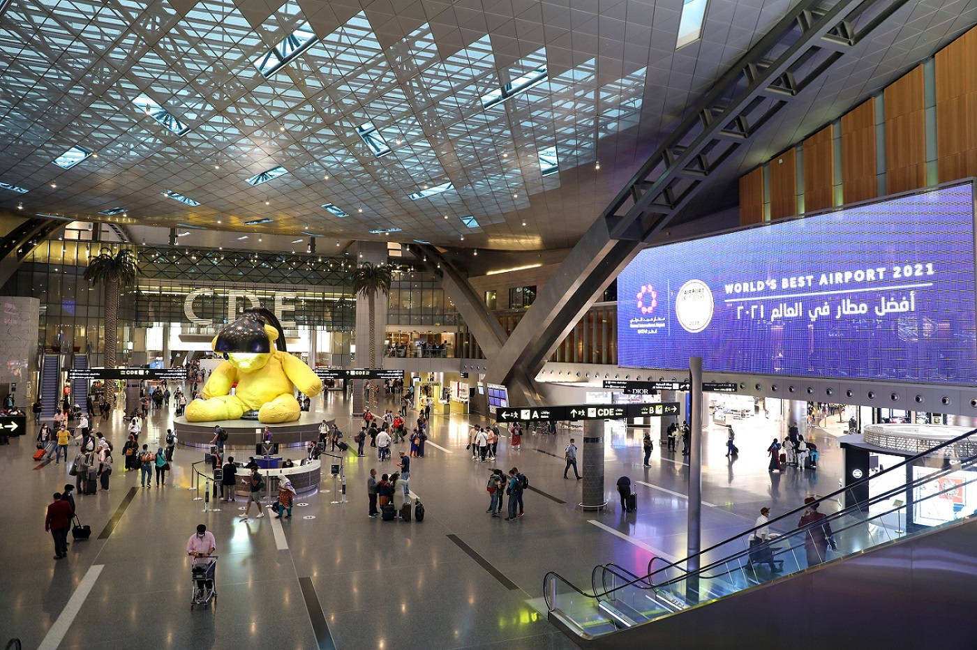 Qatar 桌布 壁紙 — 一個繁忙的現代機場航廈，有一個巨大的黃色吉祥物雕像和一個顯示「World's Best Airport 2021」的大螢幕。