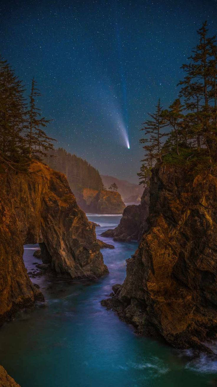 彗星劃過繁星點點的夜空，下方是岩石海岸的海灣，海水閃爍著藍光。 — iPhone 14 壁紙