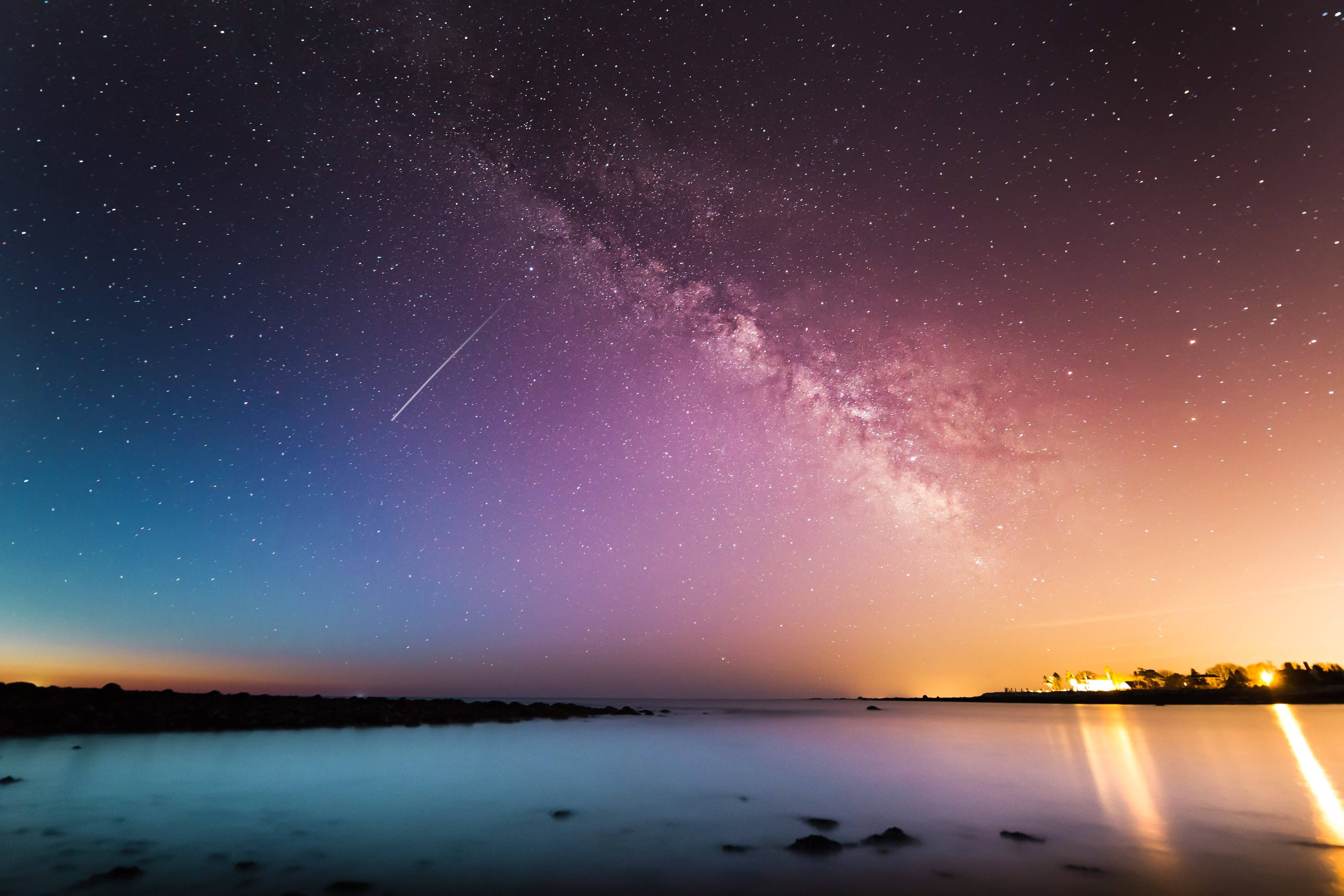 桌布 壁紙 — 一個繁星點點的夜空，一顆明亮的流星劃過平靜的湖面，湖面反射著溫暖的地平線燈光。
