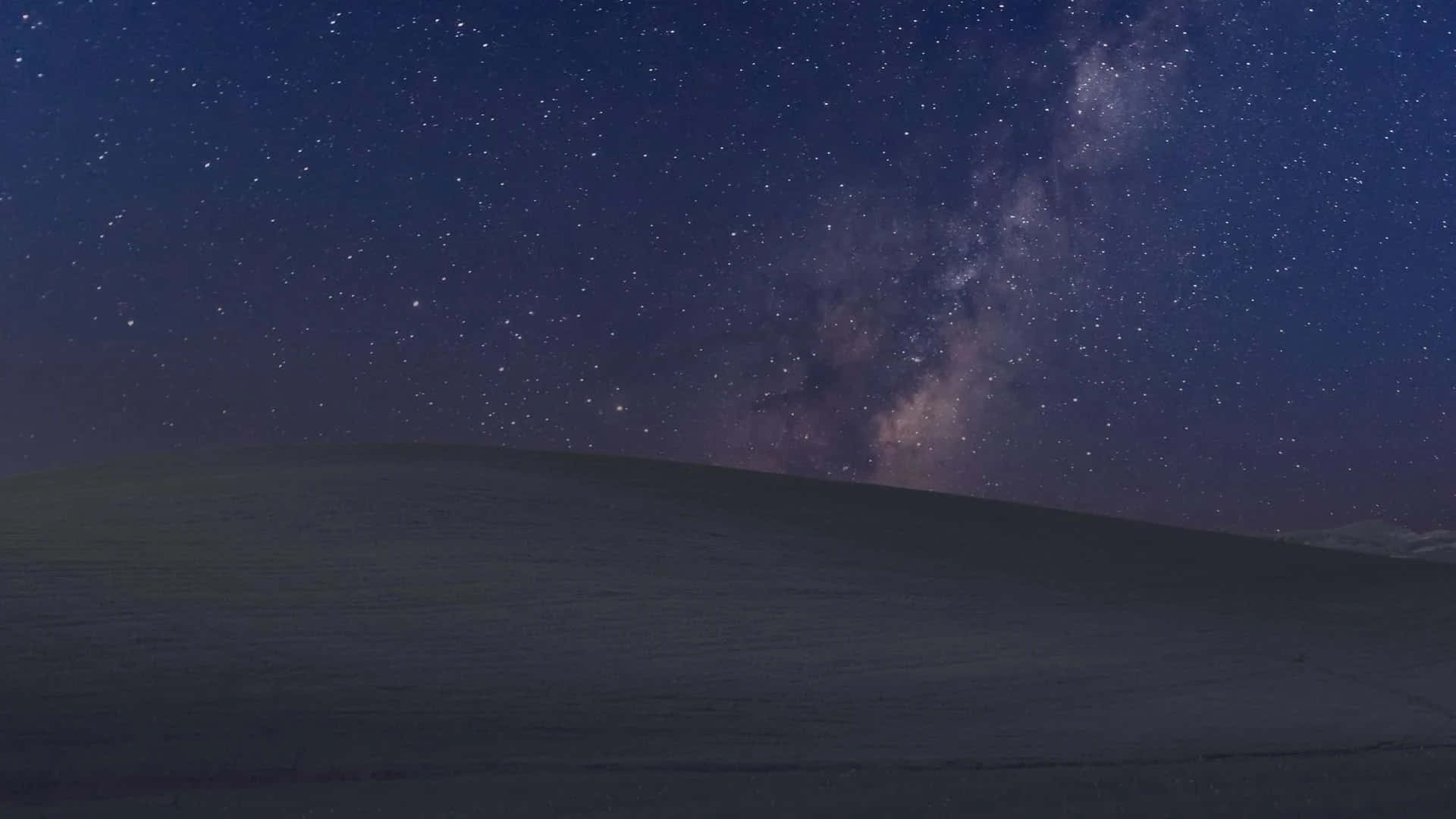 Night Mode 桌布 壁紙 — 一個繁星點點的夜空，銀河在深靛藍和紫羅蘭色調下拱起在黑暗起伏的山丘上。