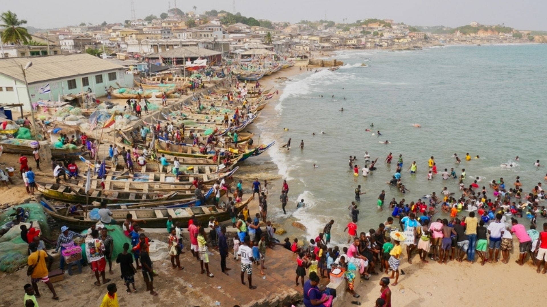 Ghana 桌布 壁紙 — 一個繁華的海岸小鎮，色彩繽紛的船隻停泊在擁擠的海灘上，人們在陰天游泳和聚集。