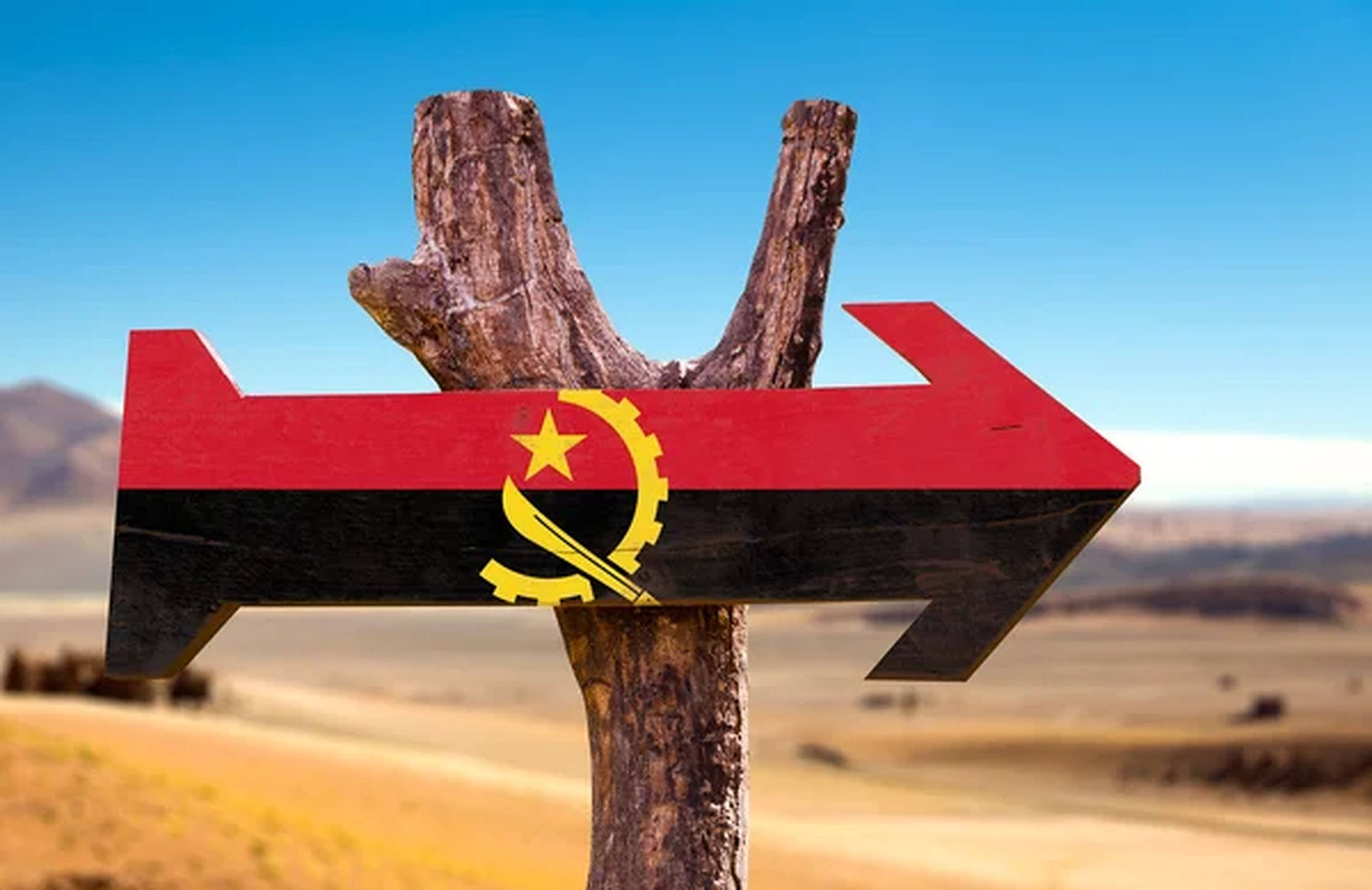 Angola 桌布 壁紙 — 一個繪有安哥拉國旗的風化木箭頭標誌，在乾燥稀樹草原上方的清澈藍天下指向右方。