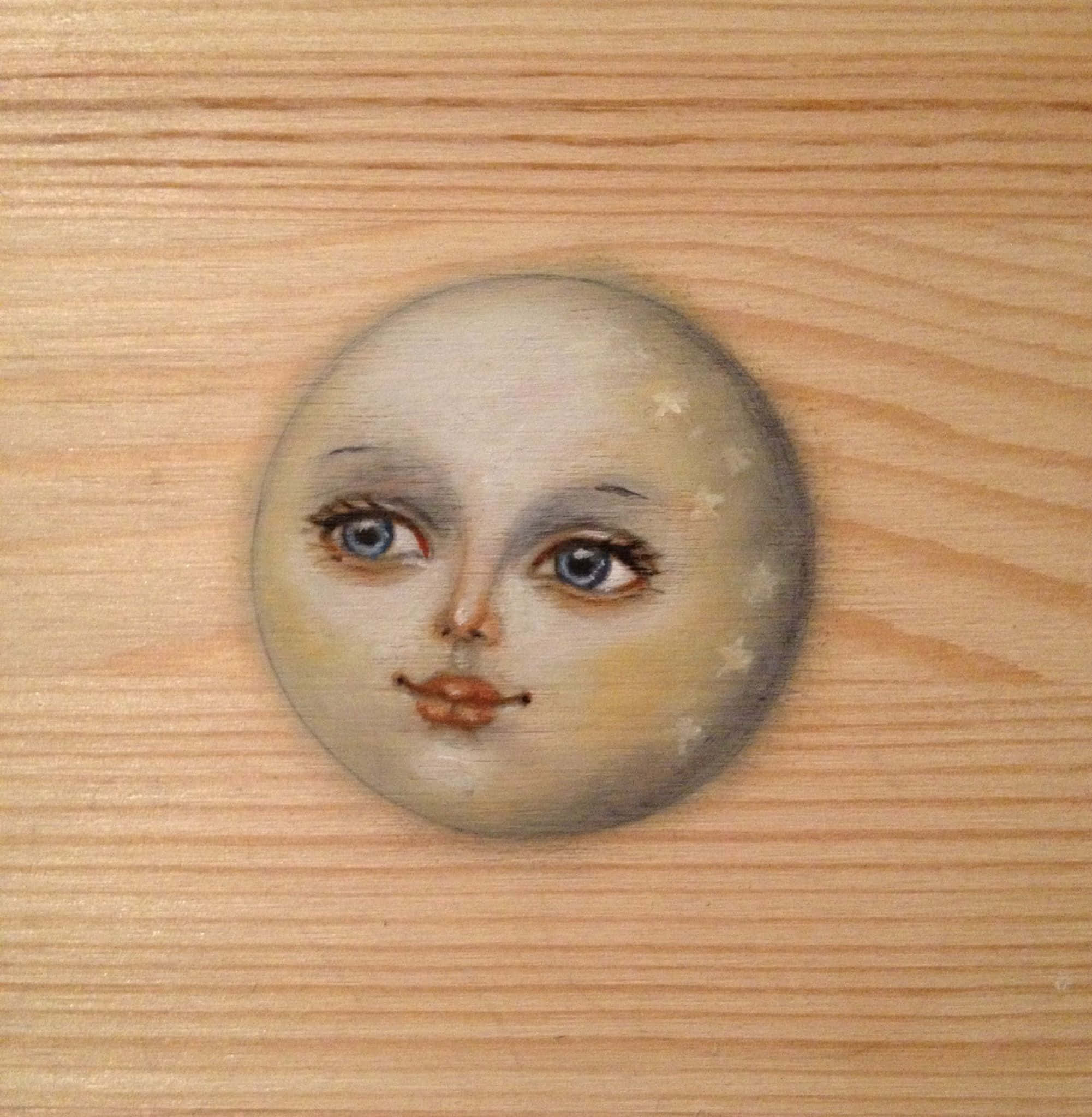 Moon Face 桌布 壁紙 — 一個繪有藍色眼睛和星星的月亮臉孔放在淺色木質表面上。