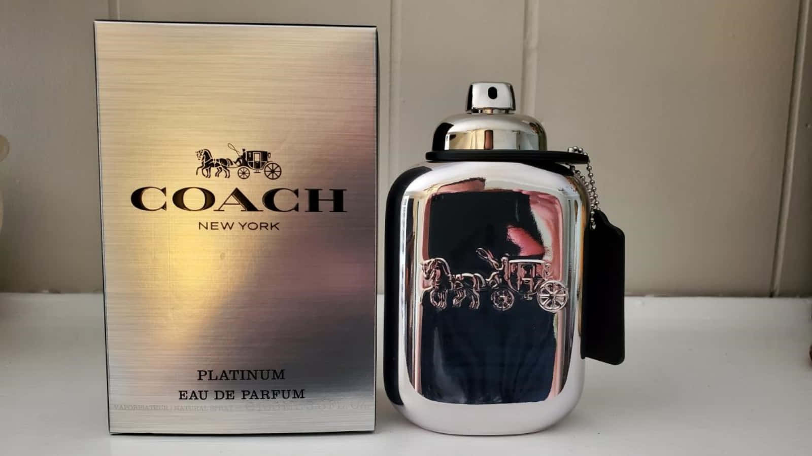 Coach 桌布 壁紙 — 一個纖細的銀色Coach Platinum Eau de Parfum香水瓶，帶有馬和馬車標誌，旁邊是其匹配的拉絲金屬盒，放在白色表面上。