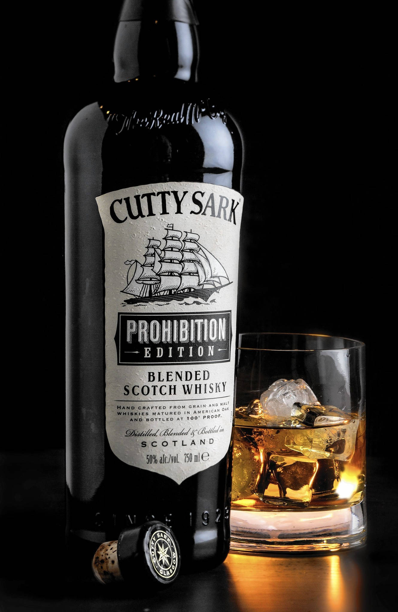 alcohol 桌布 壁紙 — 一個纖細的黑色Cutty Sark Prohibition Edition威士忌酒瓶，旁邊放著一個裝滿琥珀色液體和冰塊的岩石杯，背景為深色。